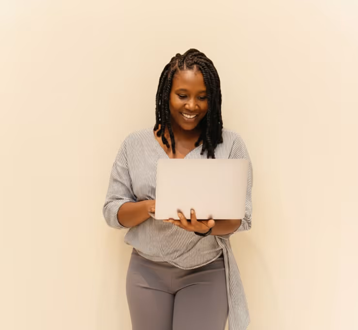 Smiling woman with braided hair standing against a beige wall using a silver laptop.