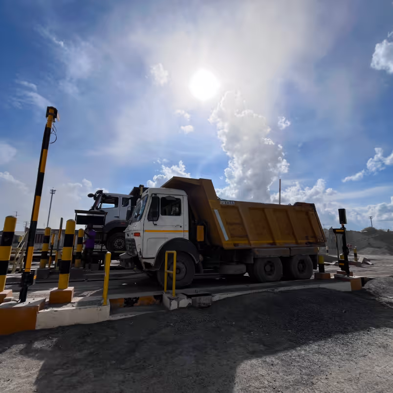 Yellow and white dump trucks at a weighbridge under a bright sun and cloudy sky.