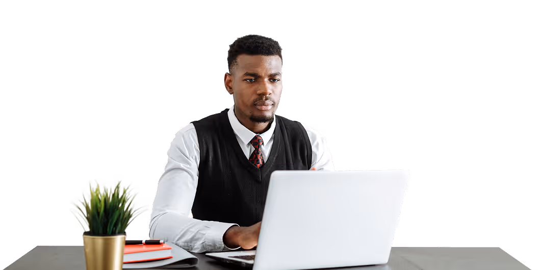 Man in white shirt and black vest focused on laptop at desk with plant and books nearby.