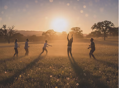 Five people playing and reaching for a ball in a sunlit grassy field at sunset.