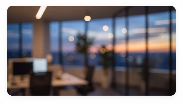 Blurred office interior with desks, chairs, computers, and large windows showing a sunset.