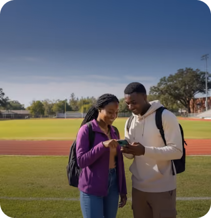 Two young adults with backpacks looking at a smartphone on a sports field under a clear sky.