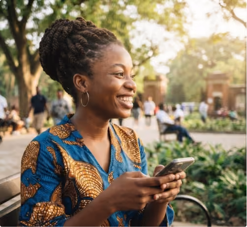 Smiling woman in patterned blue and brown shirt holding phone on a park bench with trees and people behind.