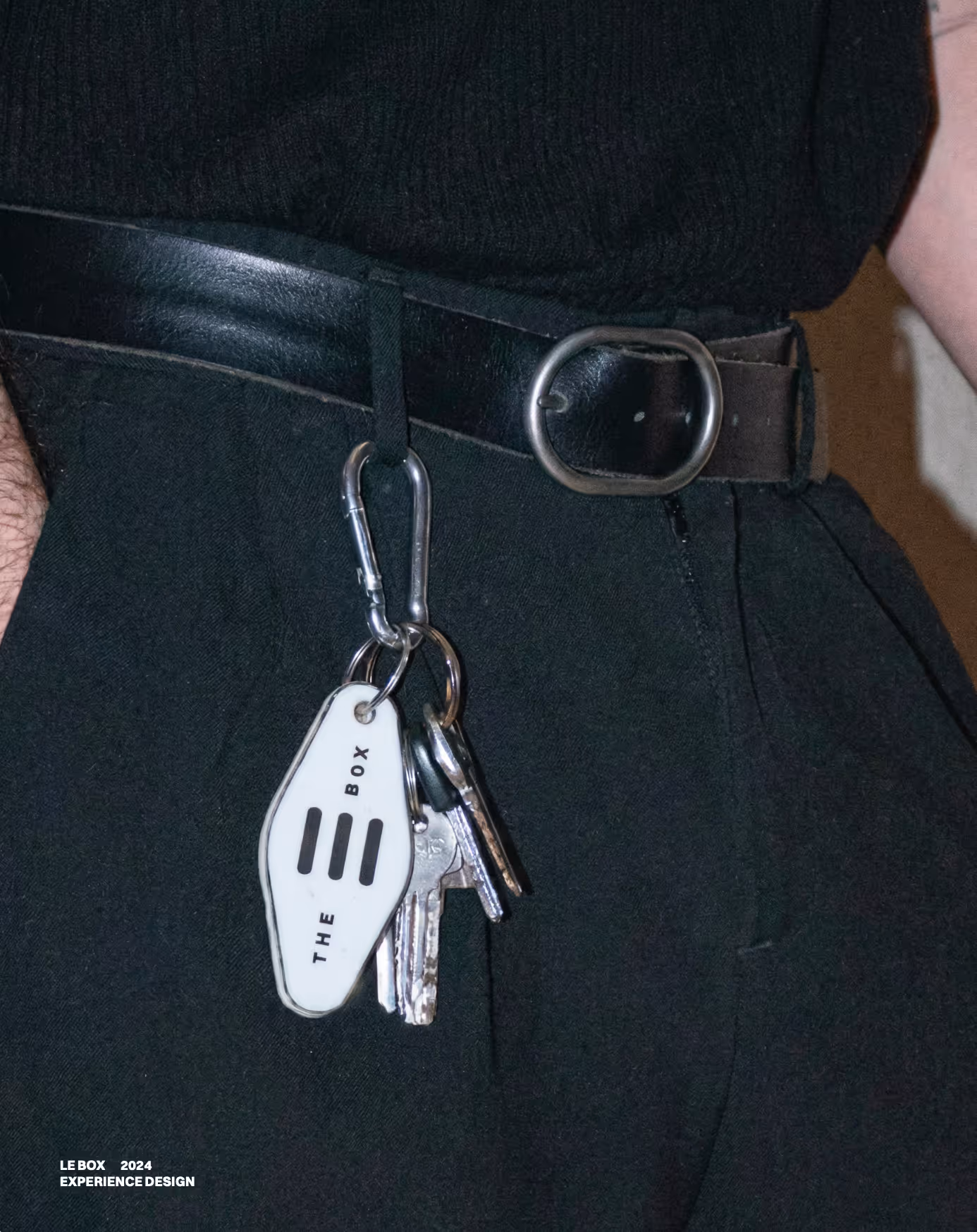 Keys hanging from a belt loop with a white keychain labeled THE BOX on a person wearing black pants.