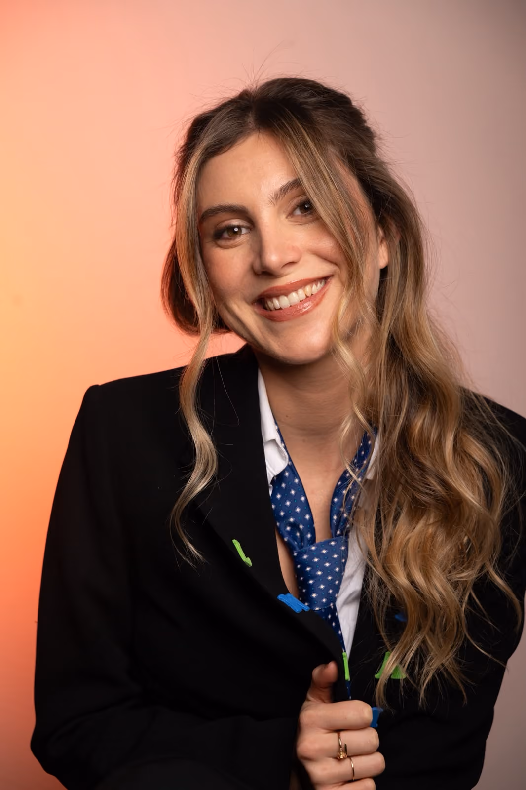 Smiling woman with long wavy hair wearing a black blazer and blue patterned tie against a warm background.