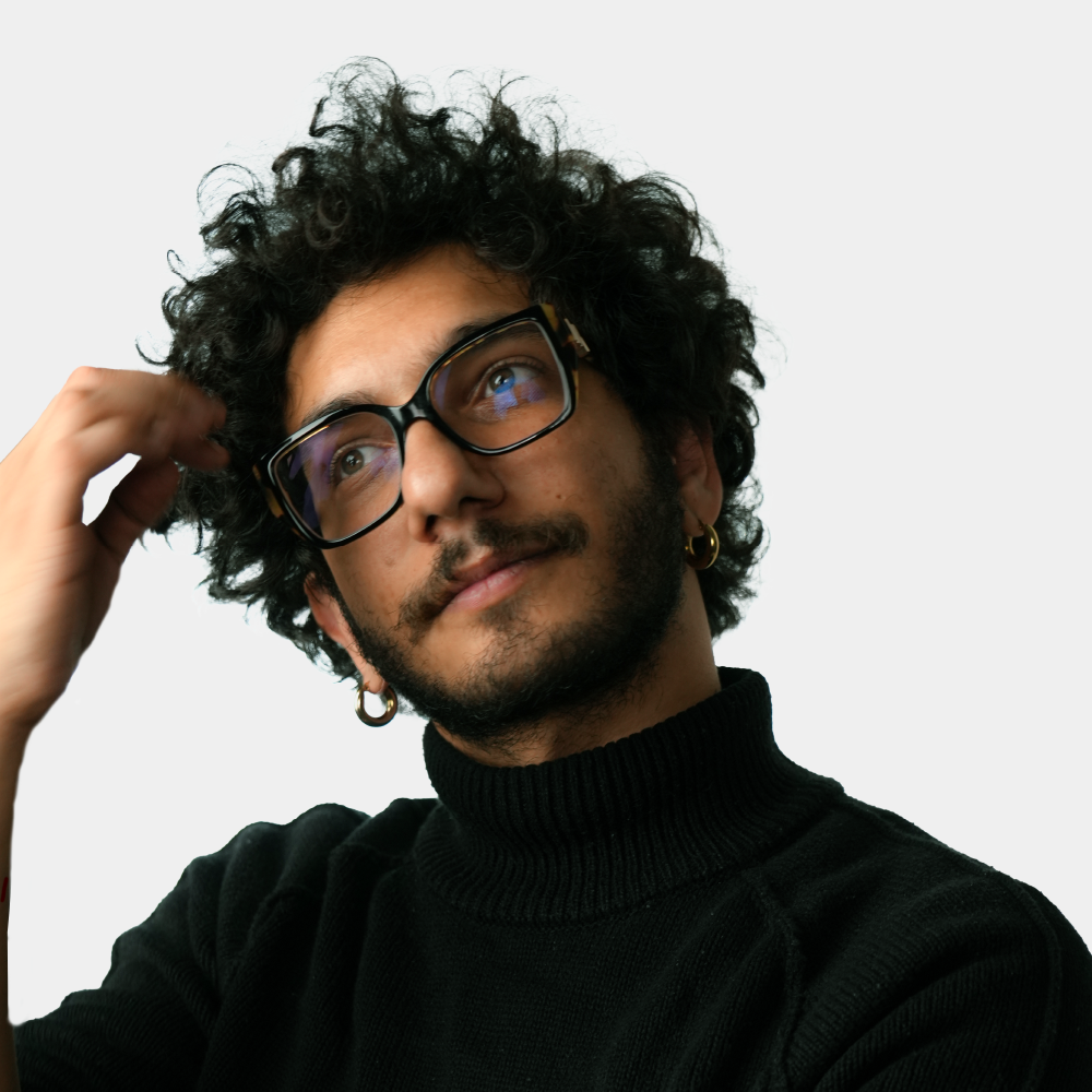 Man with curly hair and glasses wearing a black turtleneck touches his hair on a white background.