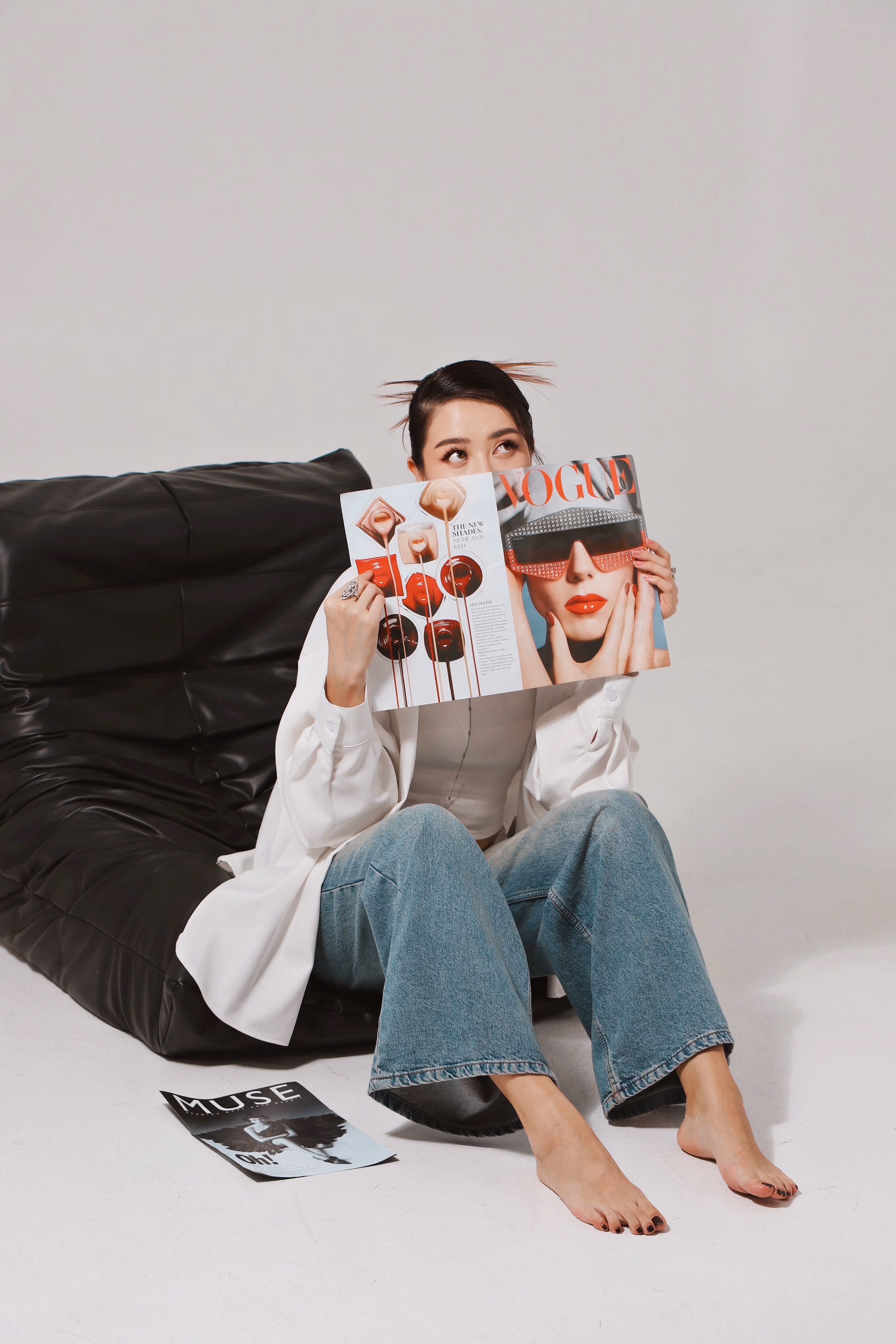Woman sitting barefoot on white floor, holding Vogue magazine partly covering her face, looking up.