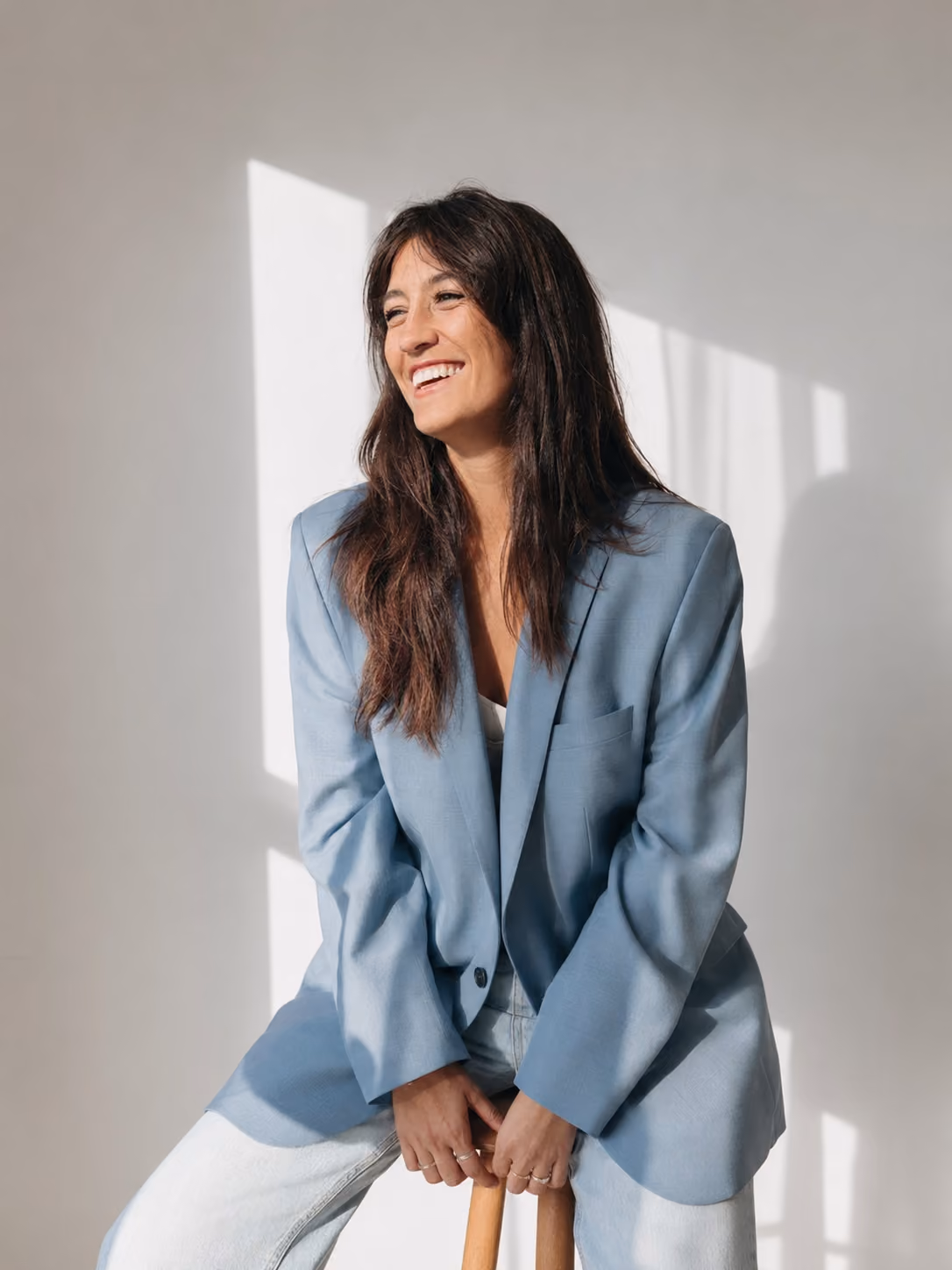 Smiling woman in light blue blazer and jeans sitting on wooden stool by window light.