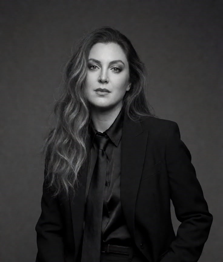 Black and white portrait of a woman with wavy hair in a black suit, shirt, and tie against a gray background.