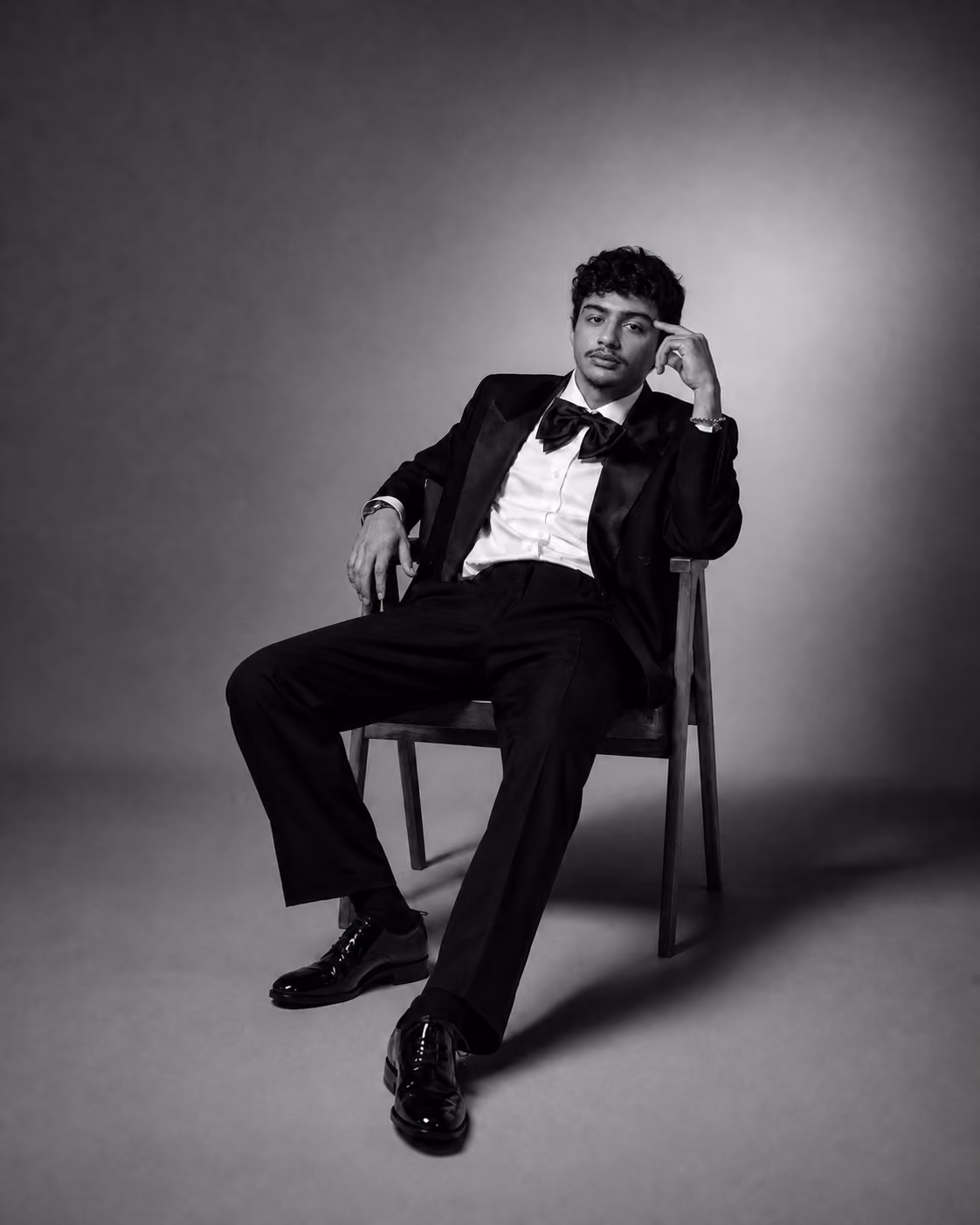 Man in a tuxedo with a bow tie sitting on a chair in a relaxed pose on a gray backdrop.