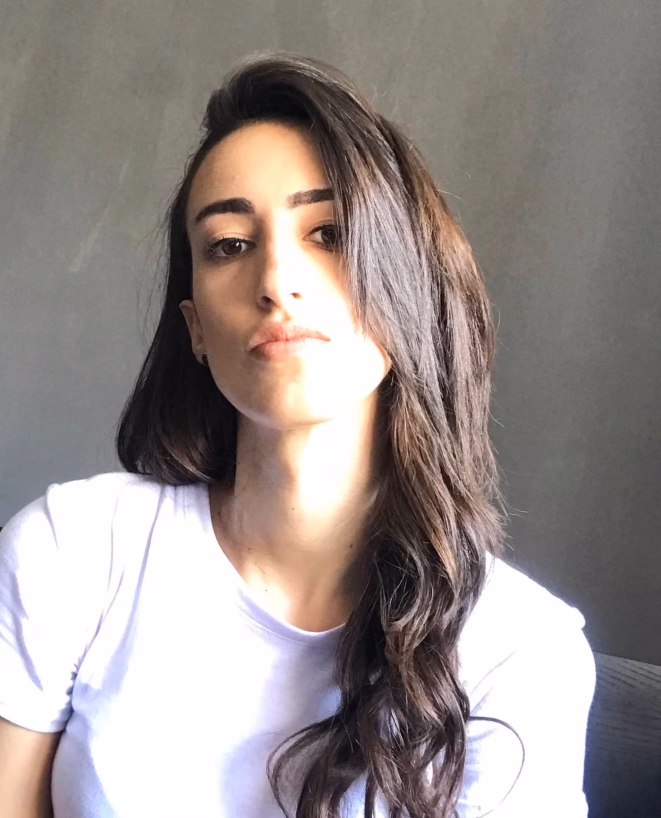 Young woman with long dark hair wearing a white shirt against a plain gray wall.