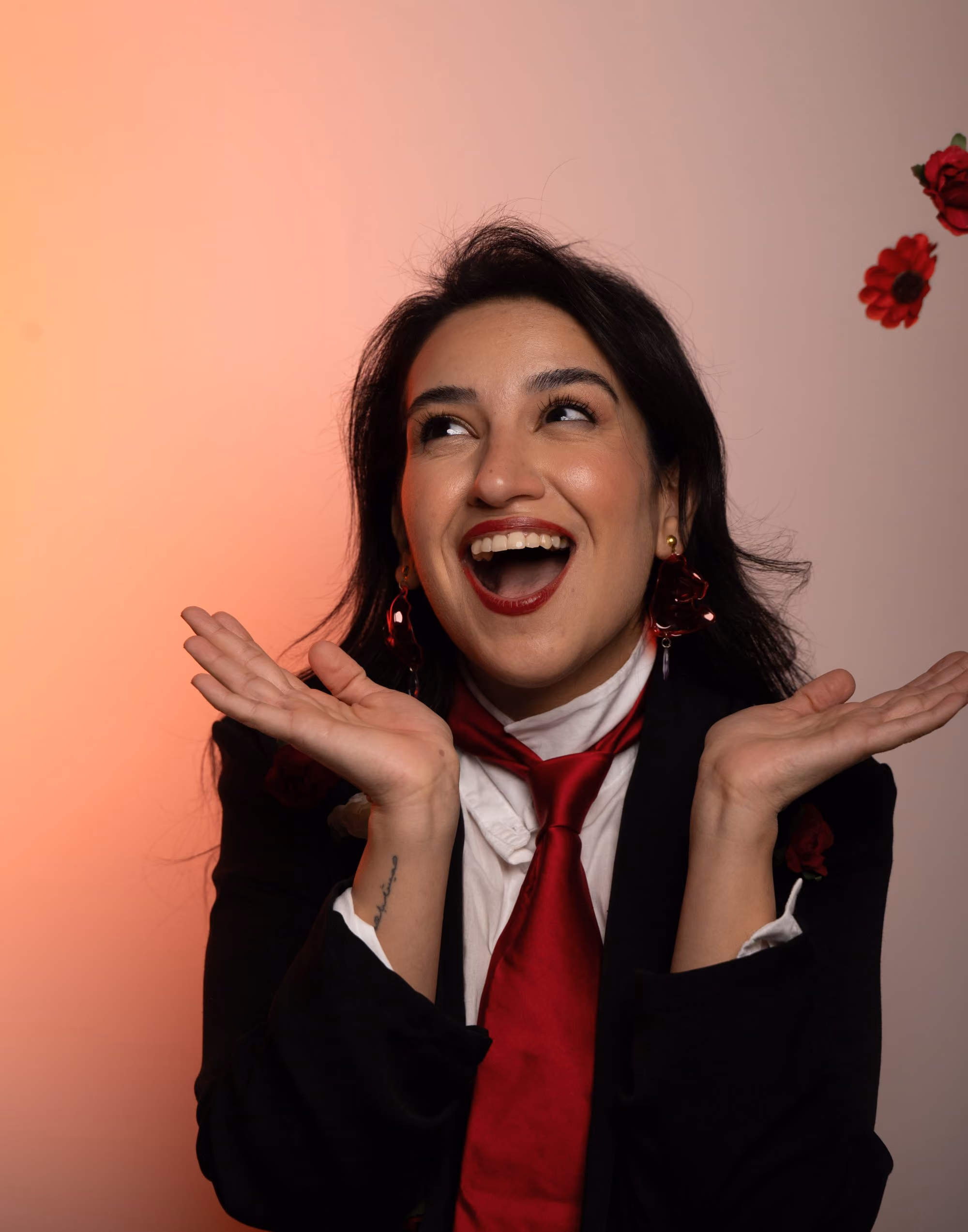 Smiling woman with black hair, red lipstick, red tie, and earrings against gradient orange background.