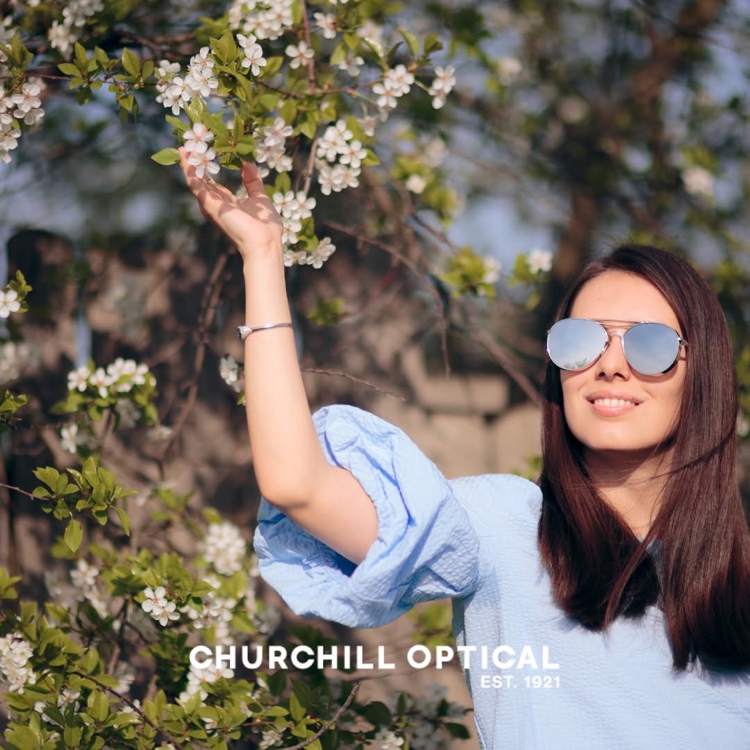Smiling woman in sunglasses and blue blouse touching white blossoms on a flowering tree branch.