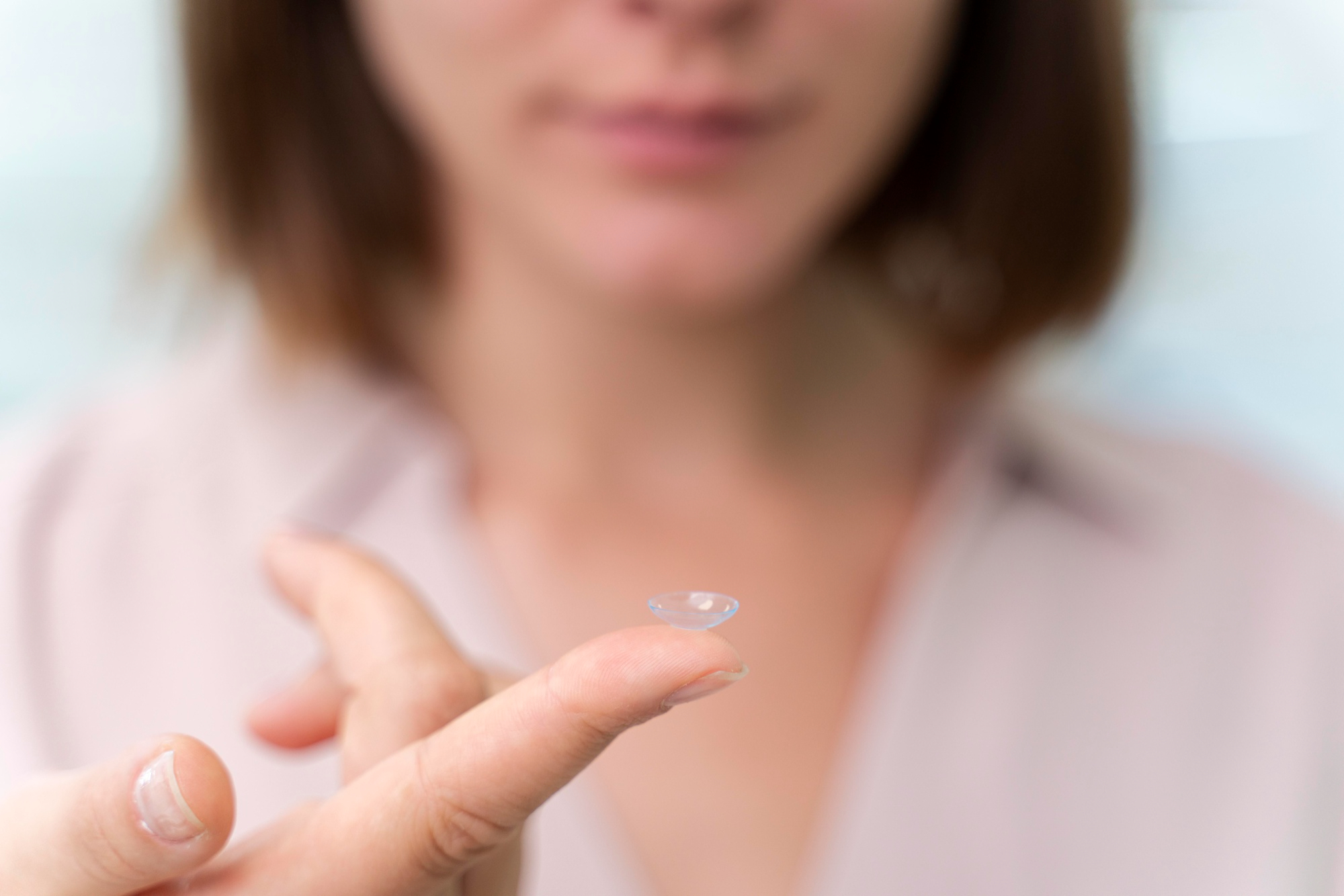 Person holding a clear contact lens on their fingertip with blurred face in the background.