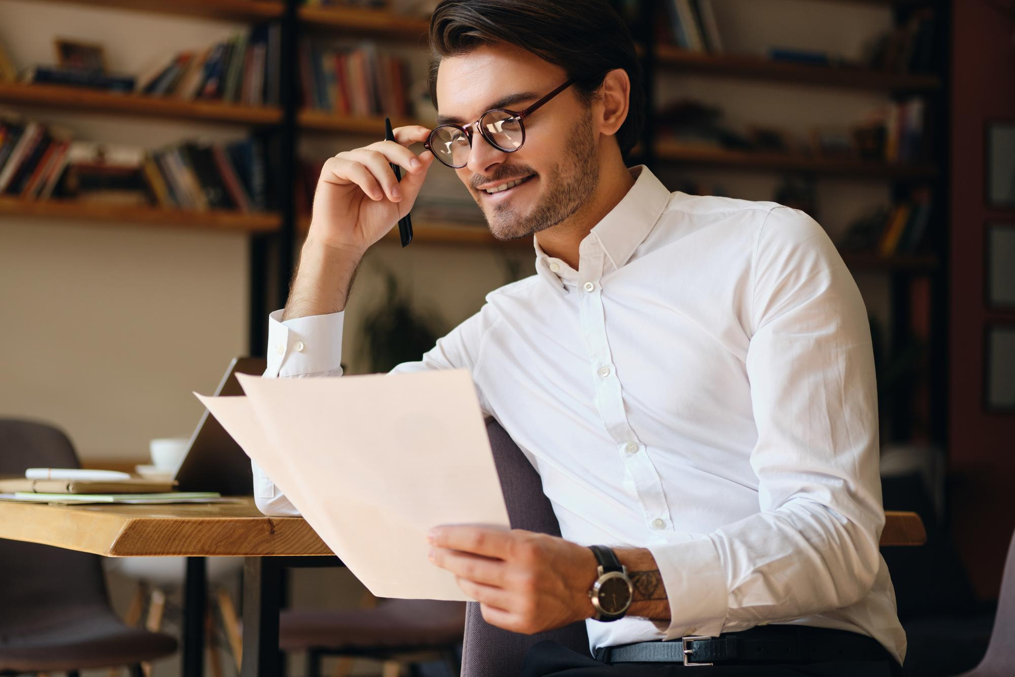 Smiling man in glasses reading papers at a table in a cozy room with bookshelves.