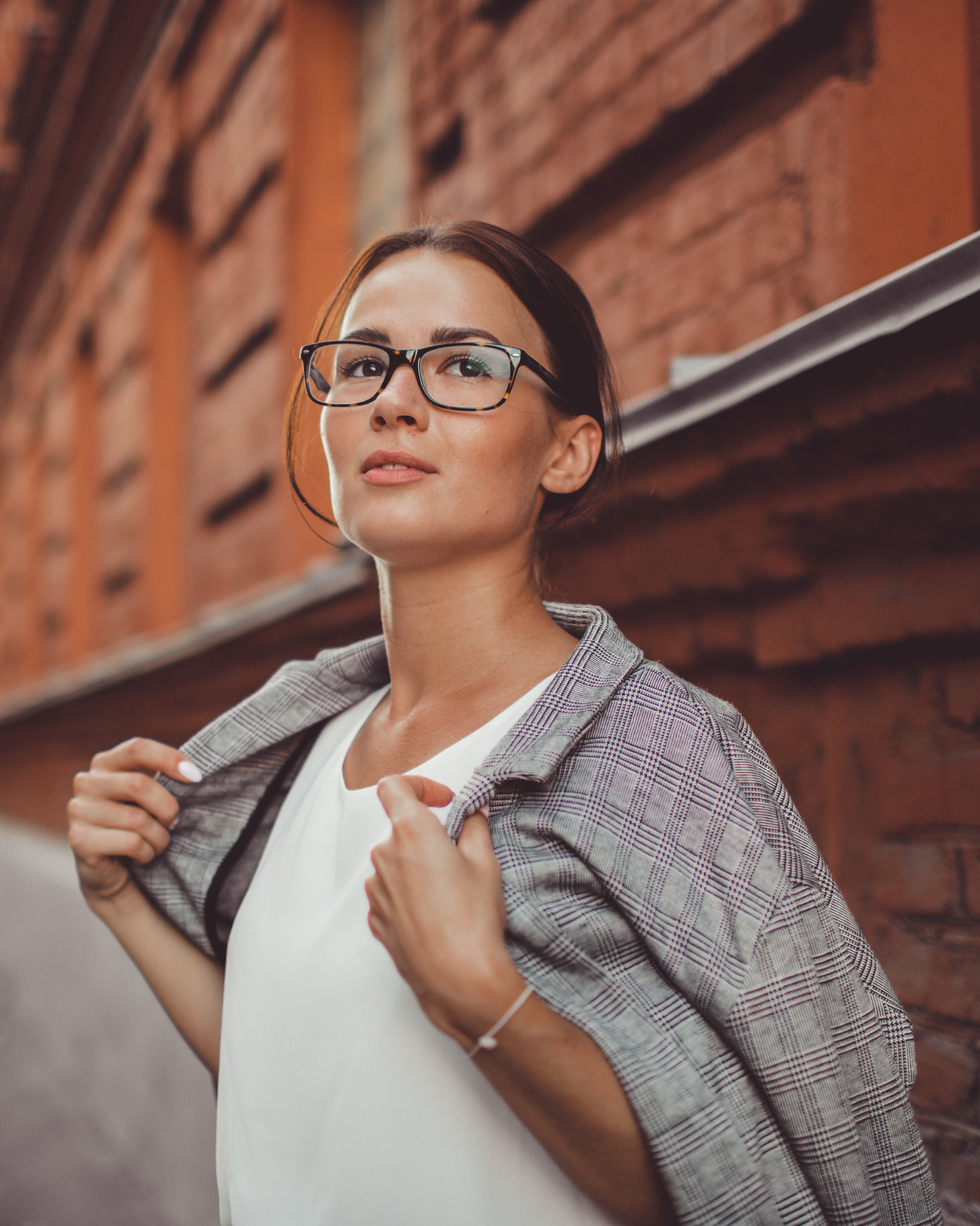 Confident woman with glasses adjusting gray plaid blazer over white top near a brick wall.
