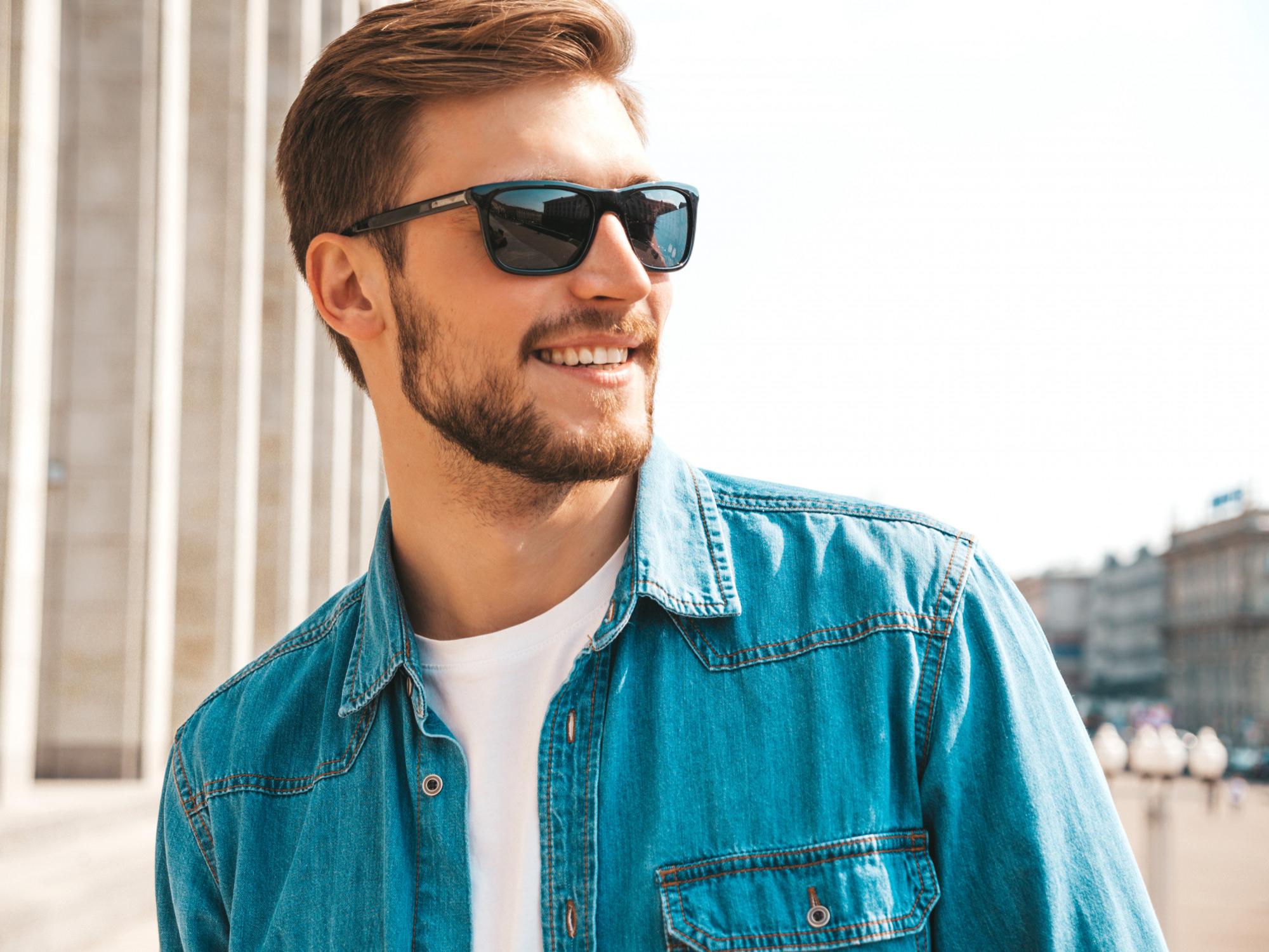 Smiling man with beard wearing sunglasses and a blue denim jacket outdoors in sunlight.