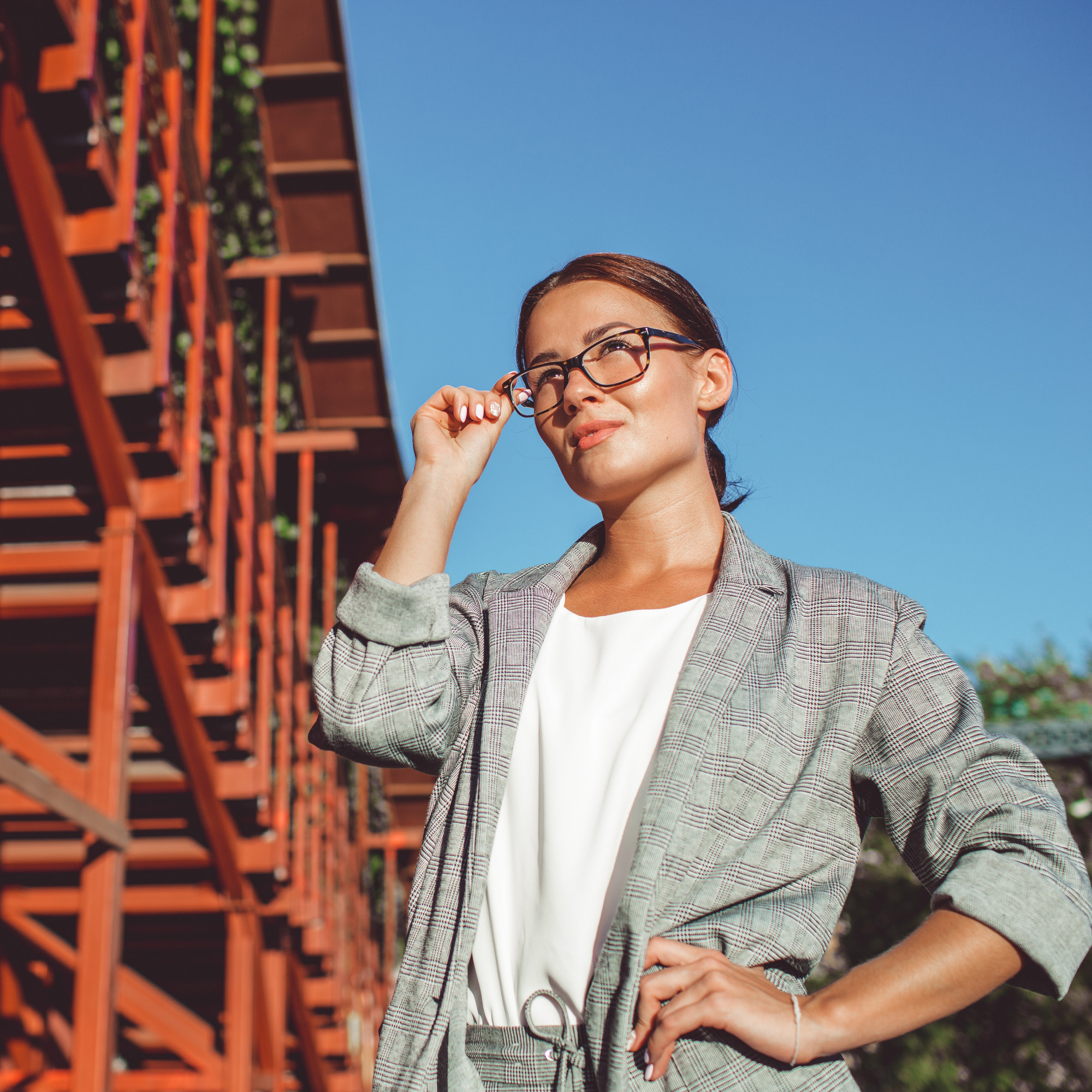Confident woman in glasses and plaid suit adjusts glasses outdoors with blue sky and red structure behind her.