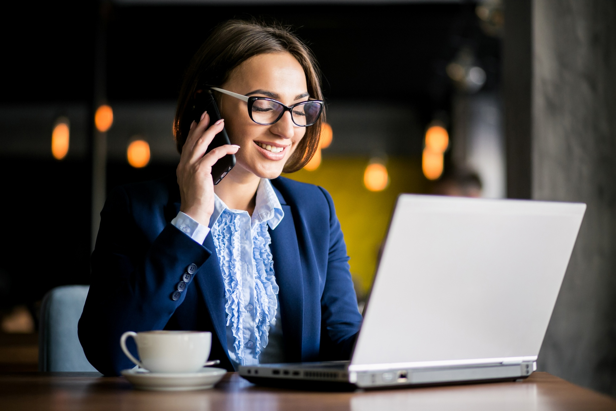 Woman with glasses in business attire smiling while talking on phone and using a laptop with coffee cup nearby.