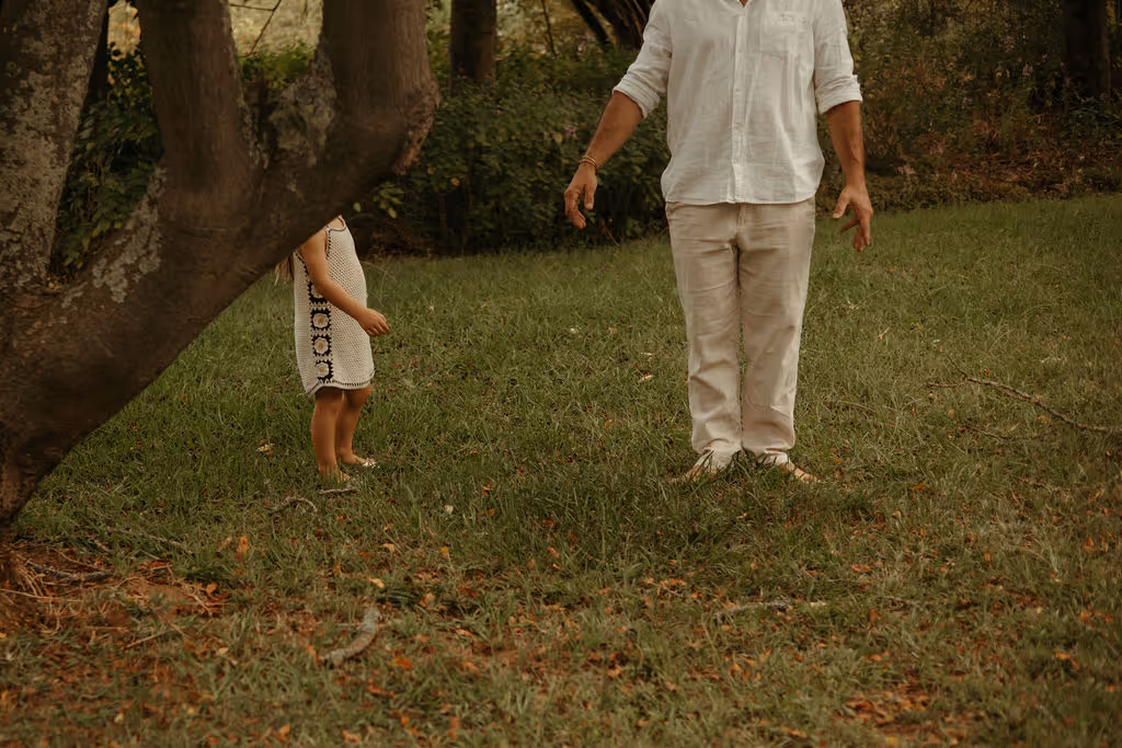 A man wearing white pants and a white shirt stands barefoot on grass beside a child in a white dress with black pattern, partially hidden behind a tree trunk in a wooded area.