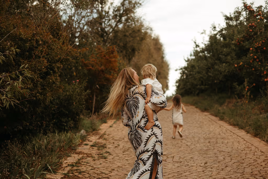 A woman with long blond hair walks down a dirt path carrying a toddler in matching patterned outfits, while a young girl in a white dress walks ahead surrounded by green trees.