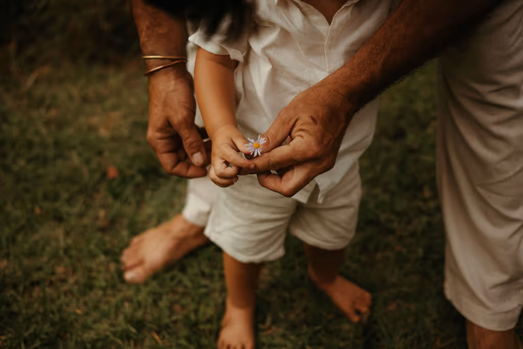 Close-up of an adult's hands gently holding a small child’s hands as the child holds a tiny daisy flower, both standing barefoot on grass.
