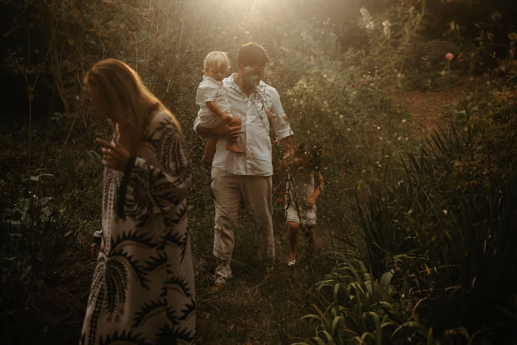 A family outdoors in a natural setting with soft sunlight filtering through foliage; a woman in a patterned dress stands in the foreground, while a man holding a young child and another child standing nearby walk in the background among plants and trees.