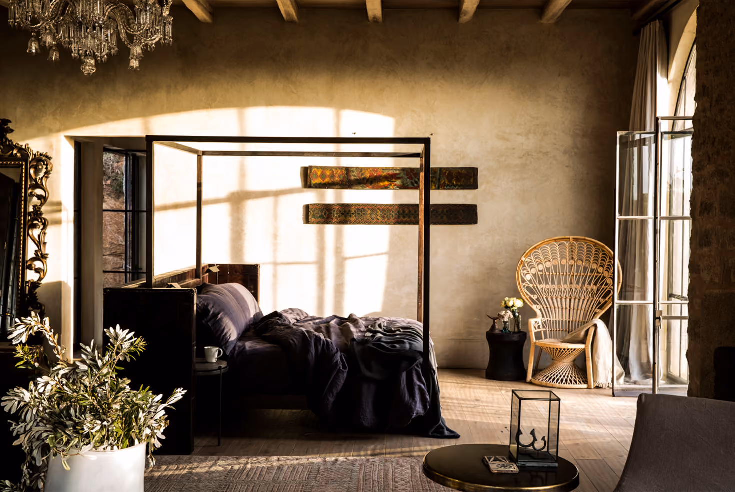 Sunlit rustic bedroom with a dark canopy bed featuring crumpled bedding, a side table with a cup, large ornate mirror, potted plant, wicker chair with a blanket, small table with flowers, and open glass doors letting in natural light.