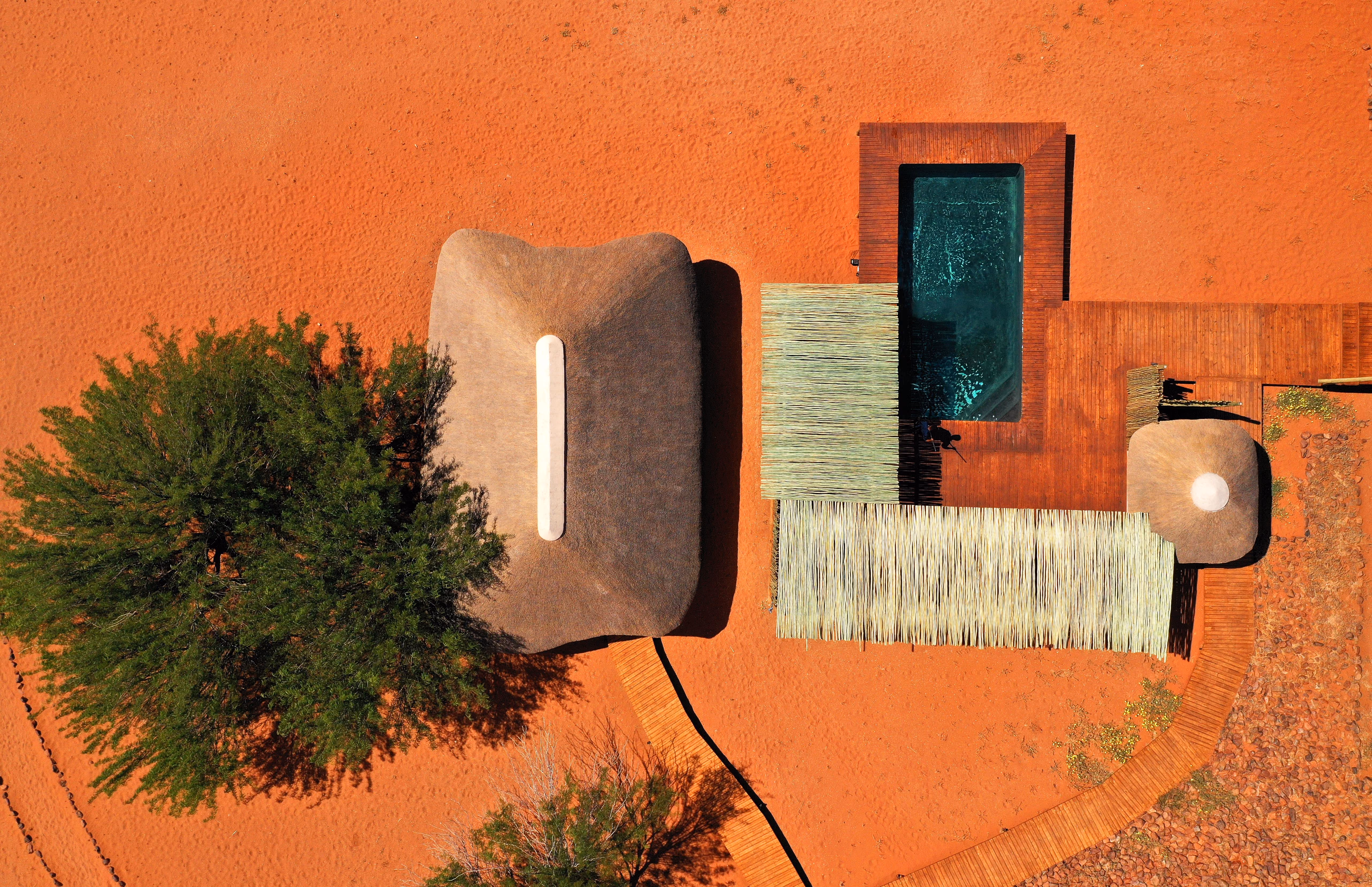 Aerial view of a desert lodge with a thatched roof building, a large green tree casting shadows on orange sand, a rectangular swimming pool with wooden decking, and a wooden walkway curving through the sand.