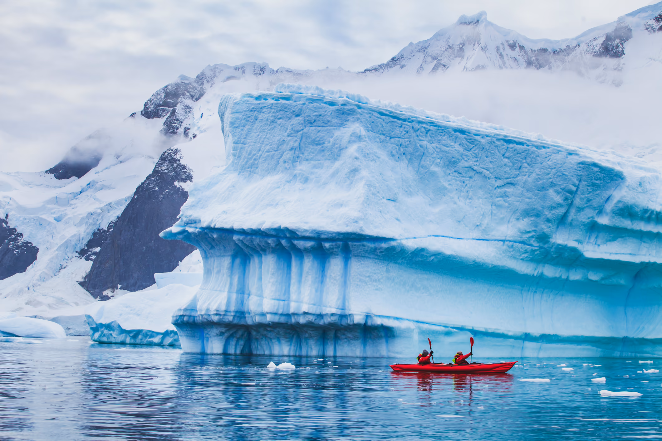 Two people in red kayaks paddling near a large blue and white iceberg in calm icy waters with snow-covered mountains in the background.