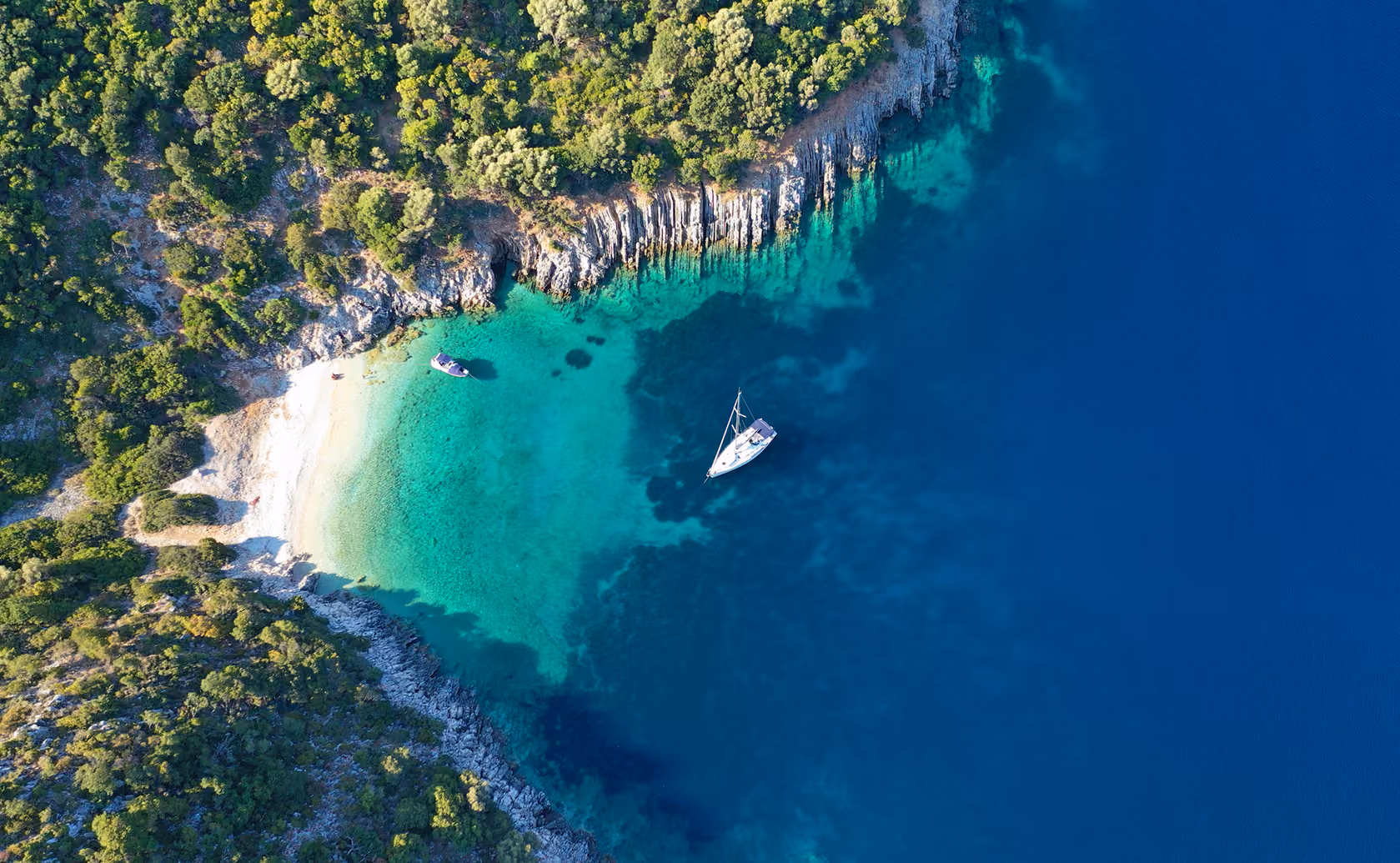 Aerial view of a small rocky island with dense green vegetation surrounding a turquoise lagoon with two boats anchored near a white sandy beach, bordered by deep blue sea.