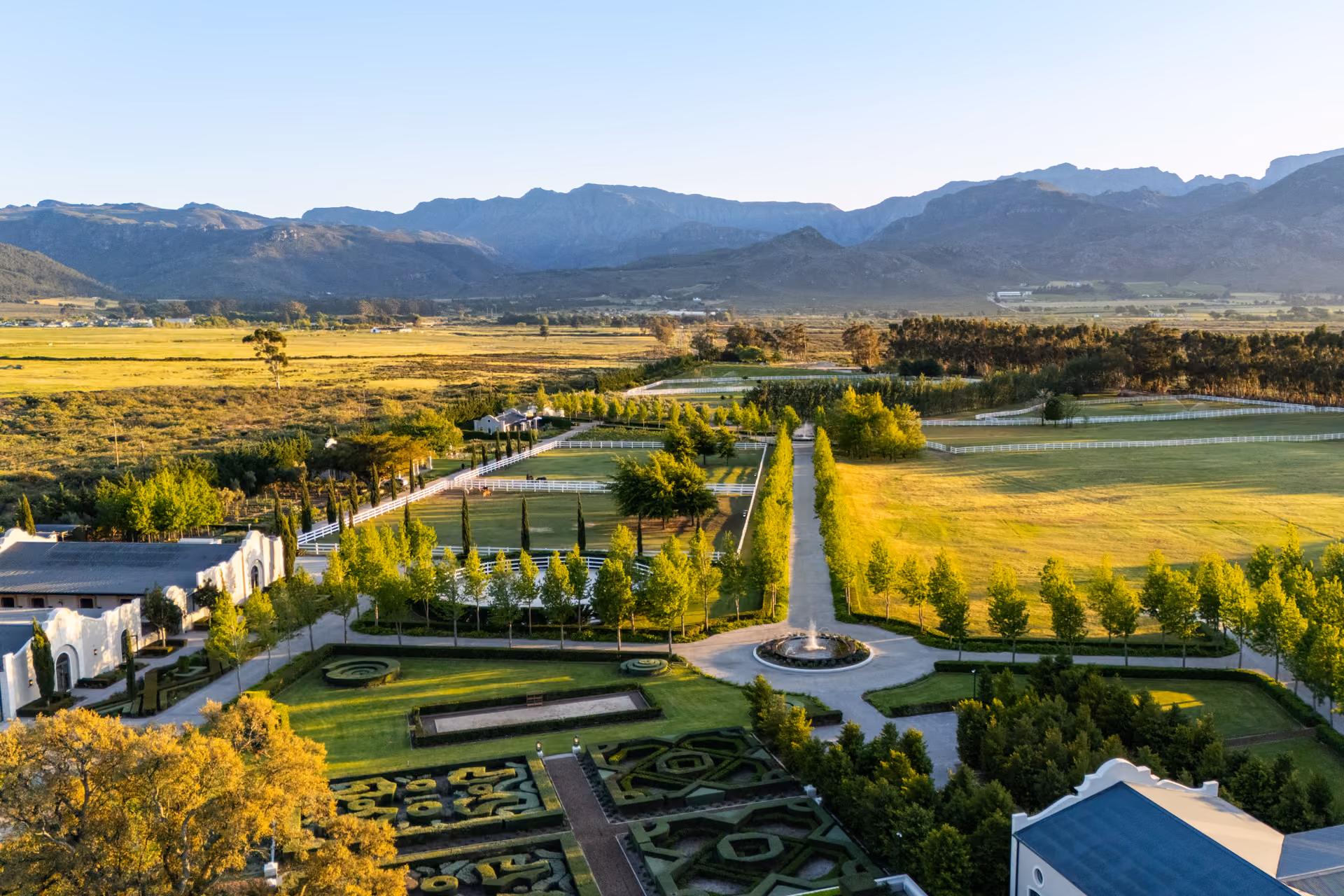Aerial view of a landscaped estate with manicured gardens, a circular fountain, tree-lined pathways, fenced pastures, and mountains in the background under a clear sky.