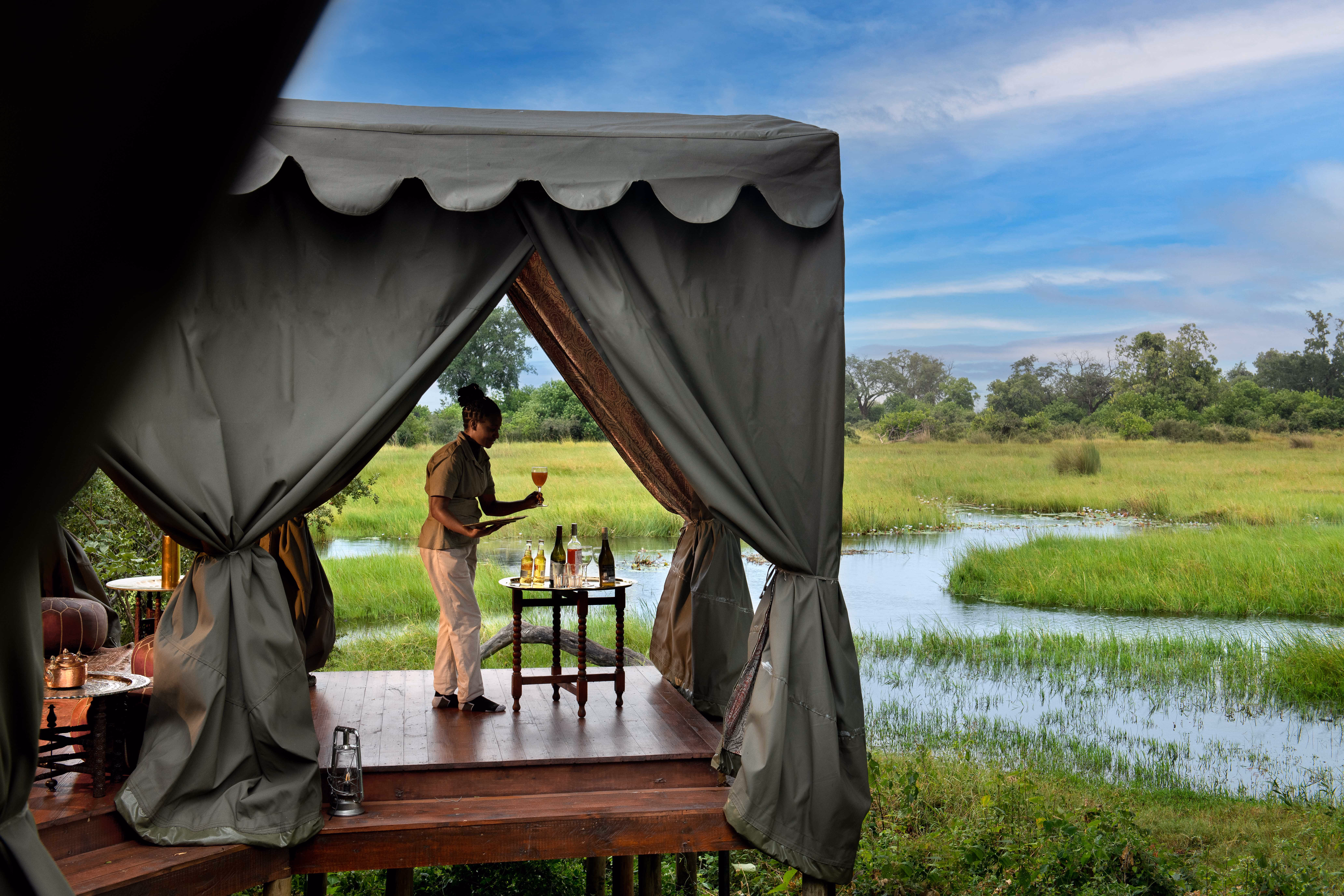 A woman standing on a wooden deck under a green canopy in a grassy wetland setting, holding a tray with a glass of drink next to a small table with bottles and glasses.