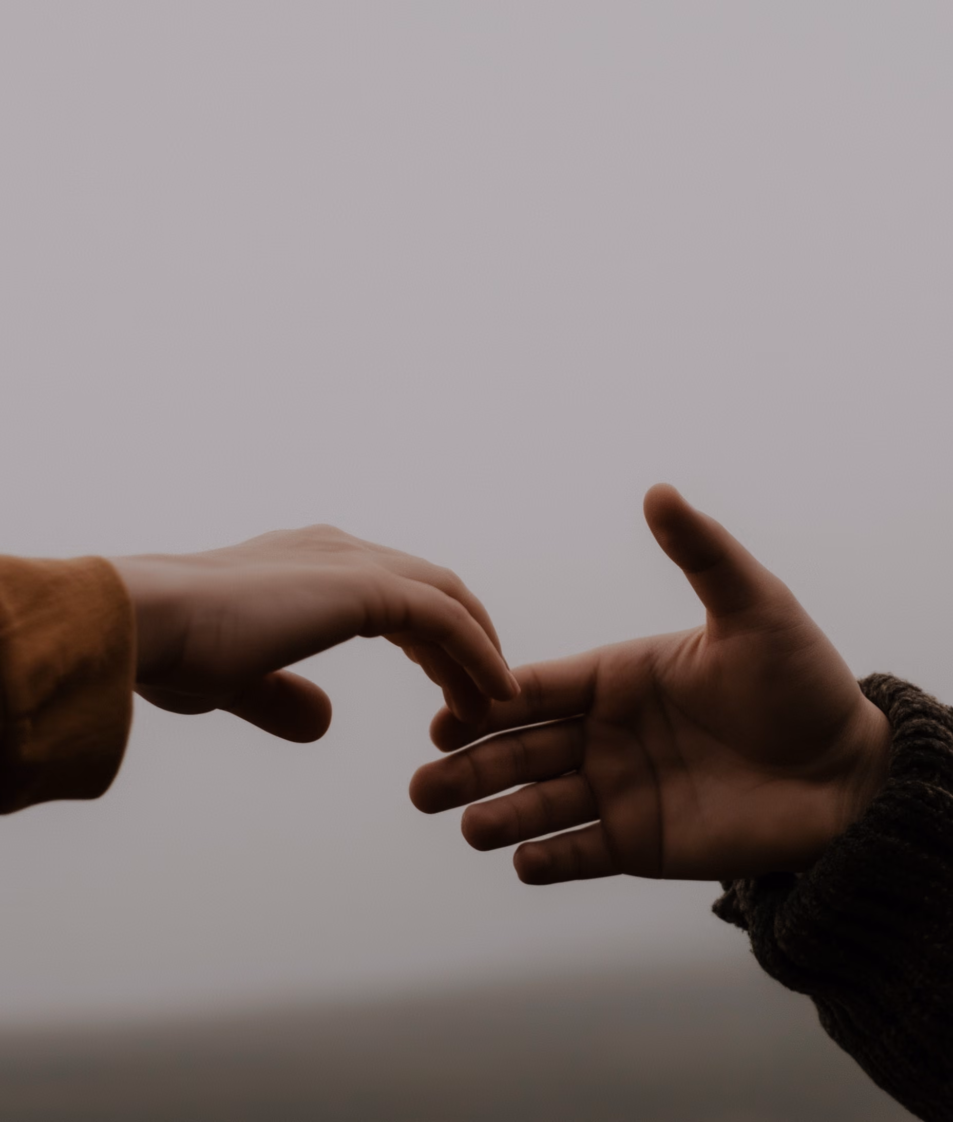 Two hands reaching toward each other against a muted, foggy background, one hand wearing a brown sleeve and the other a dark knitted sleeve.