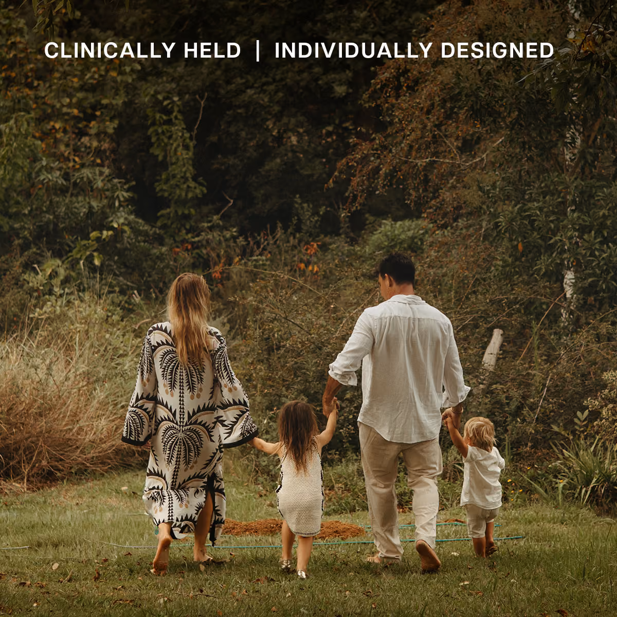 A family of four holding hands and walking outdoors on grass toward dense trees and bushes, with the text 'CLINICALLY HELD | INDIVIDUALLY DESIGNED' above them.