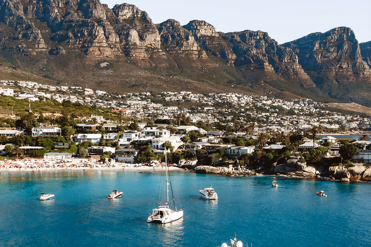 Clear turquoise bay with several boats and a small sandy beach lined with umbrellas, backed by residential houses and large rugged mountains under a clear sky.