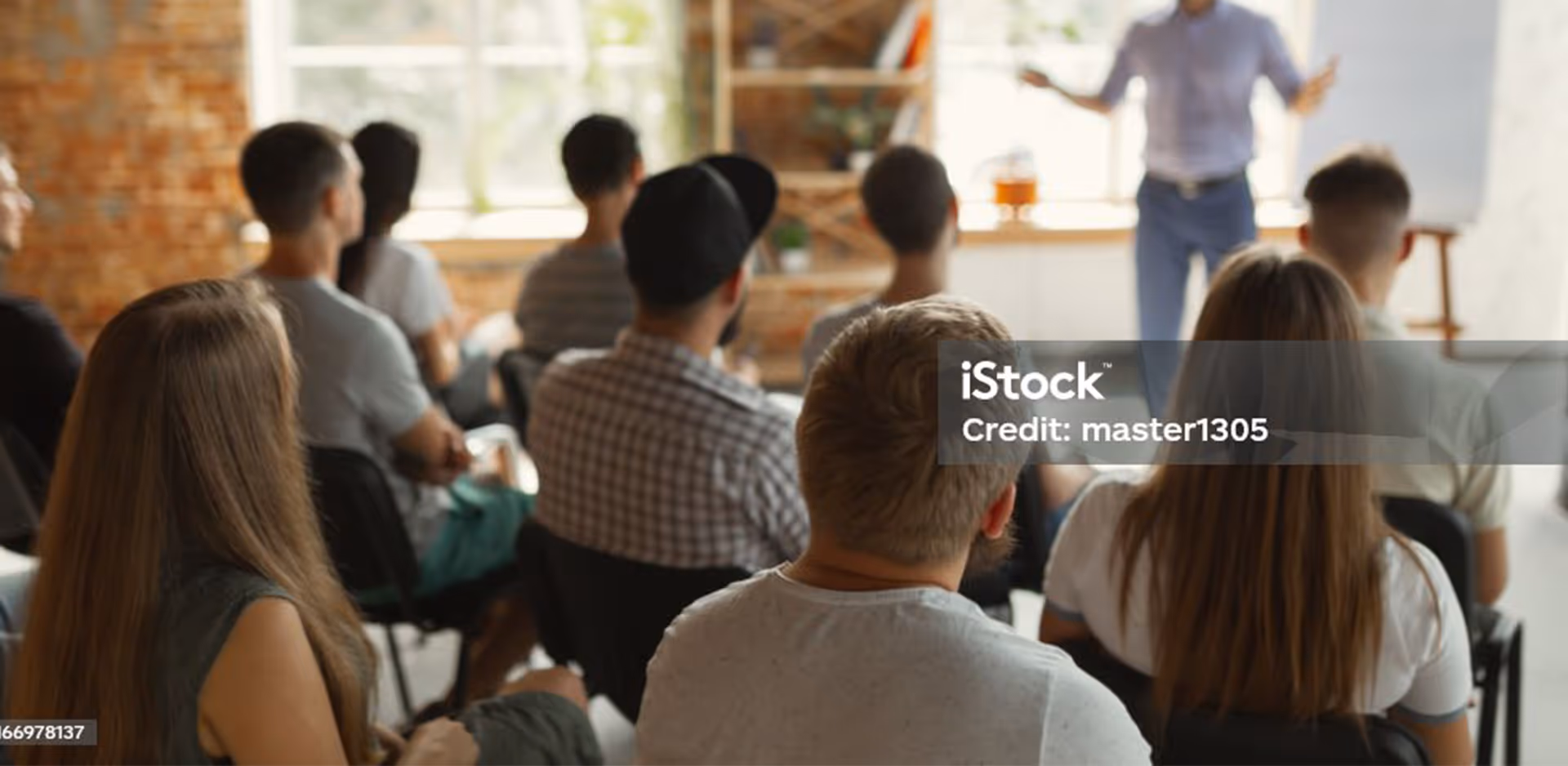 Gruppe von jungen Erwachsenen in einem Klassenzimmer, die einem Lehrer zuhören, der vor einem Whiteboard steht.