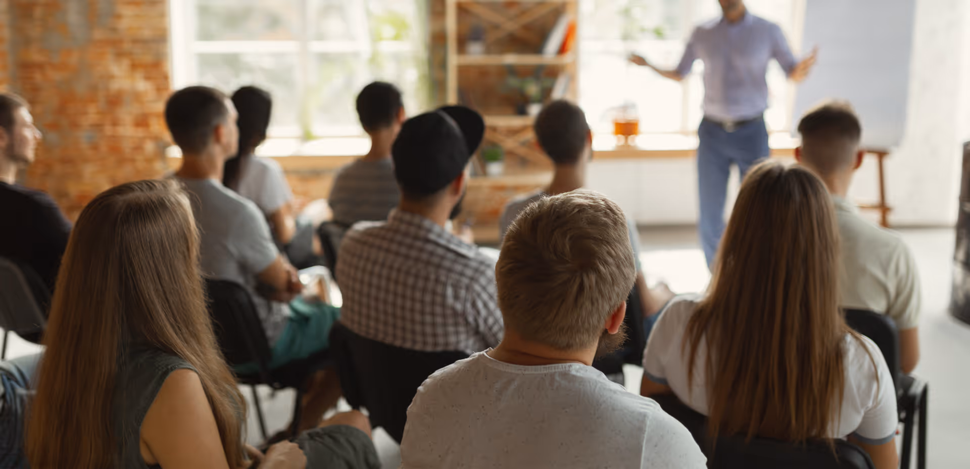 Gruppe von Menschen hört einem Präsentator in einem hellen Raum mit Backsteinwand und Fenster zu.