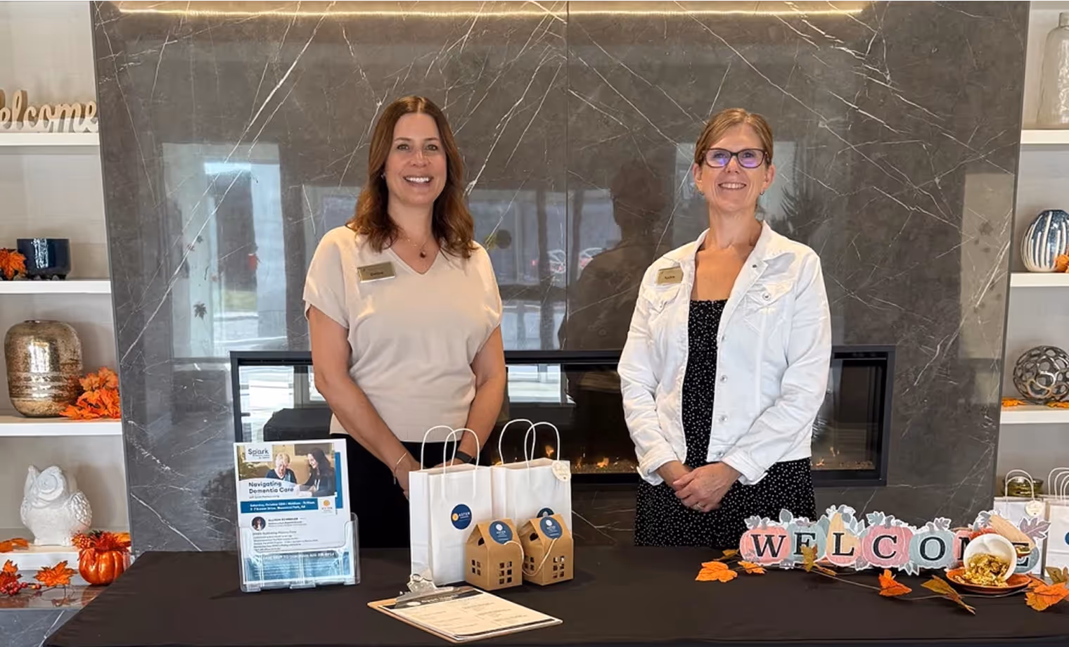Two women standing behind a table with welcome sign, gift bags, and informational materials in front of a marble fireplace.