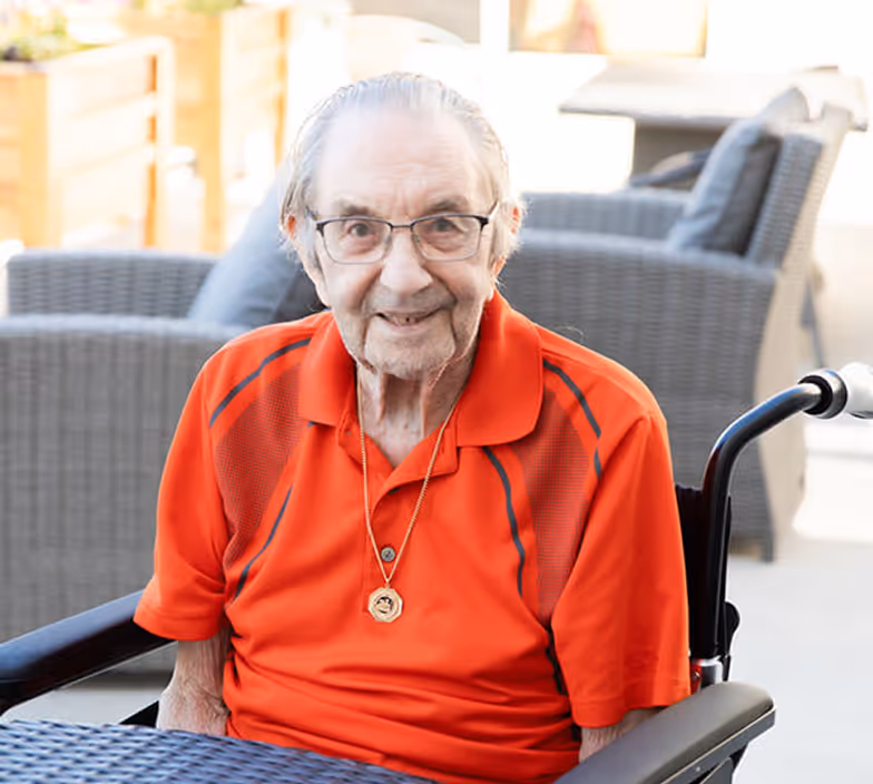 Elderly man wearing glasses and an orange polo shirt sitting in a wheelchair outdoors.