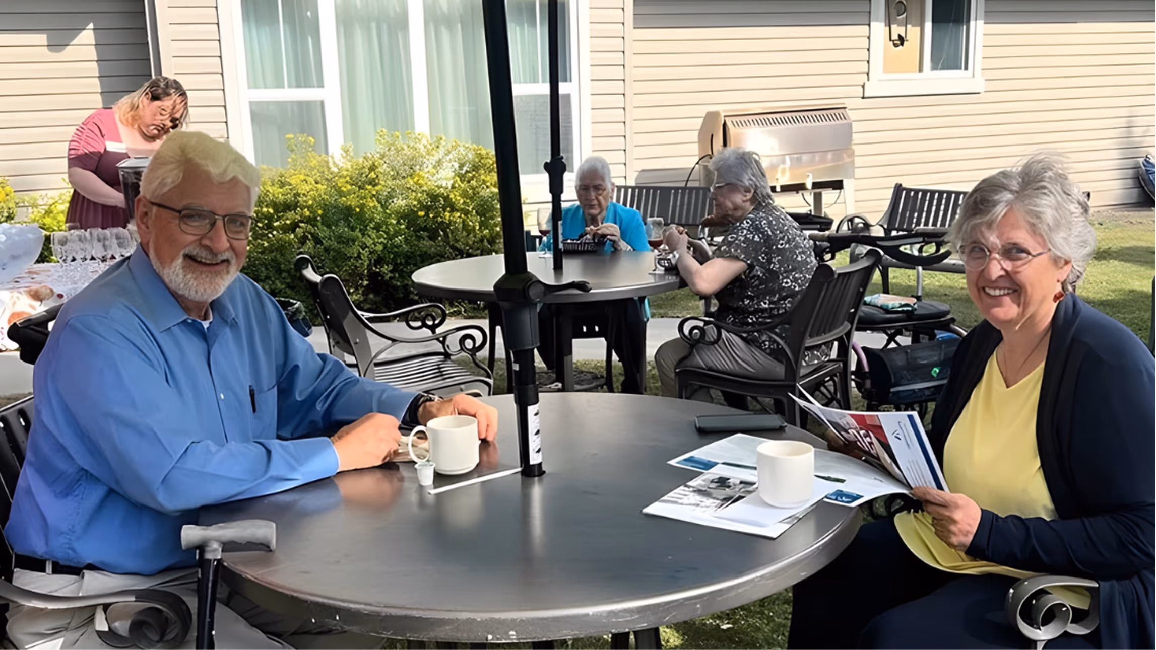 Two elderly people smiling and sitting at an outdoor table with coffee mugs, with three other elderly people sitting at a table in the background.