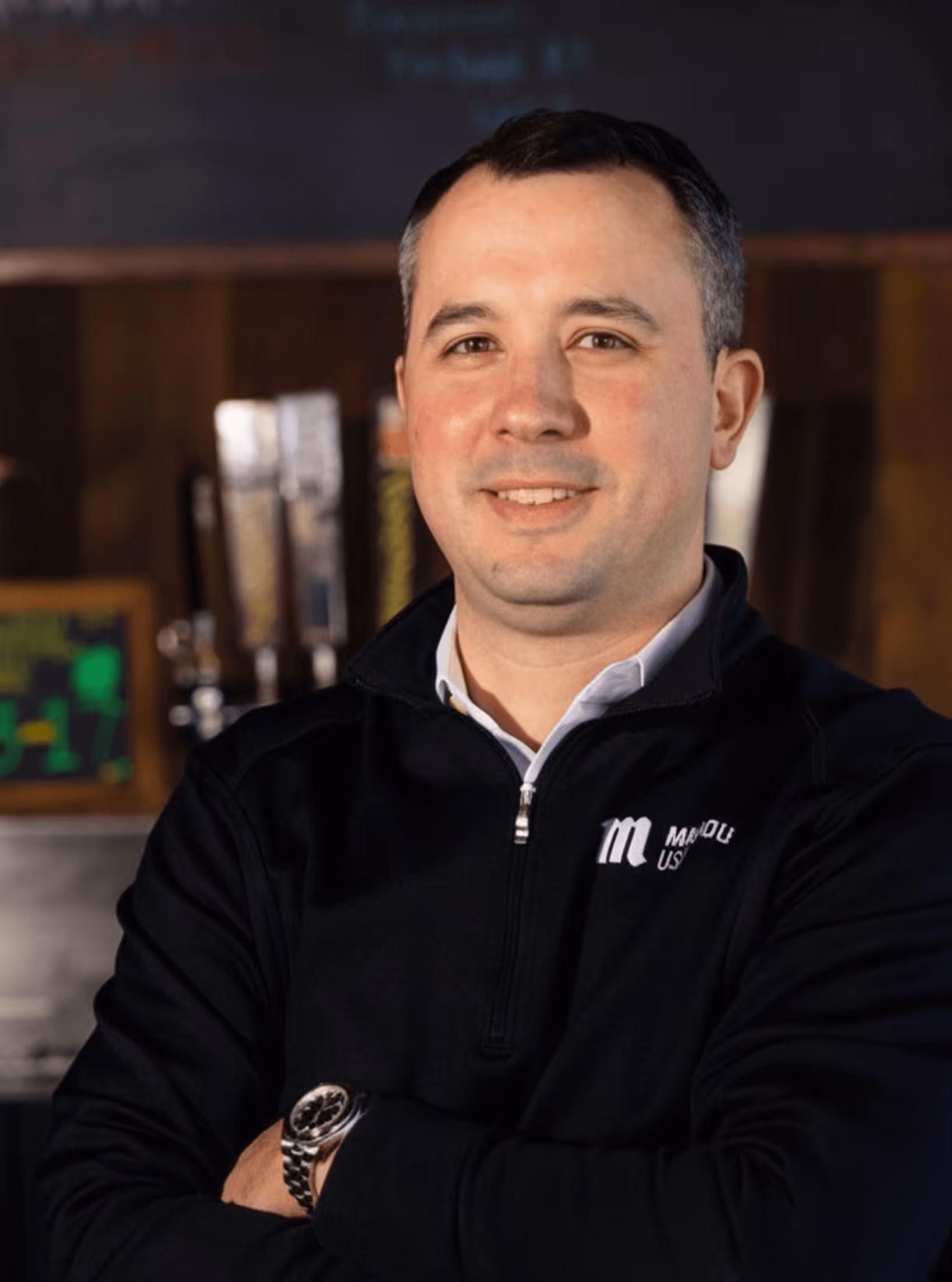 Smiling man with short dark hair wearing a black zip-up jacket and silver watch, standing with arms crossed.