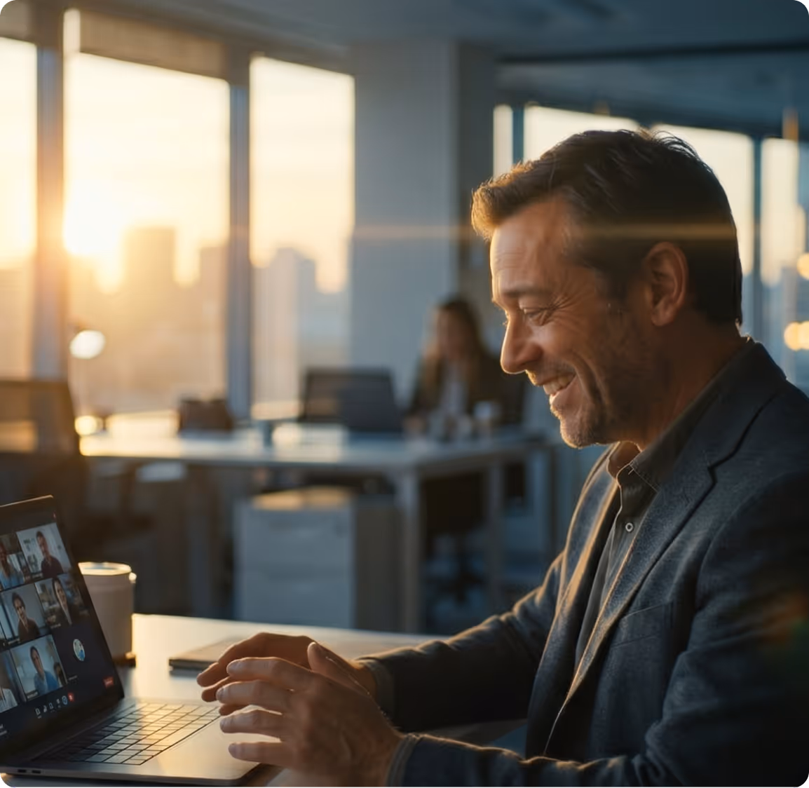Man smiling and participating in a video conference on a laptop in a modern office with sunset light.