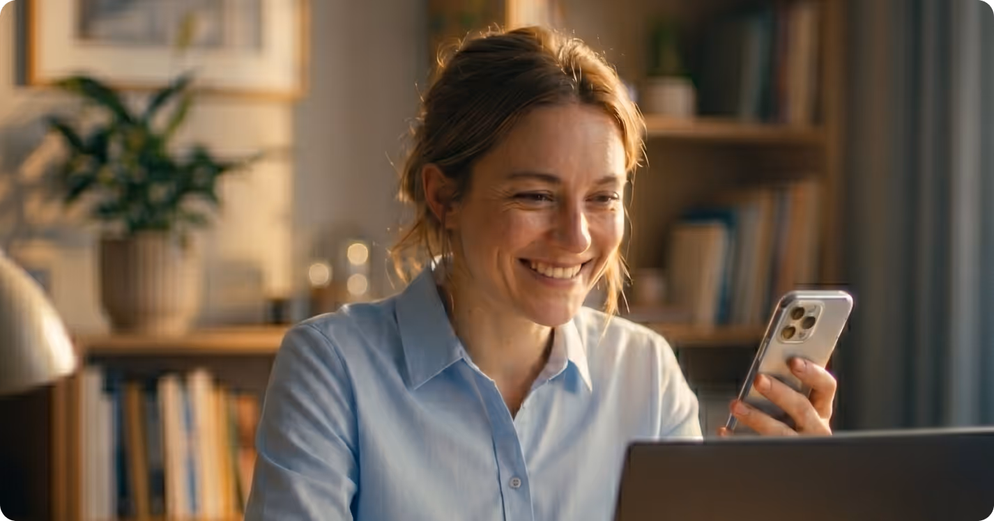 Smiling woman holding a smartphone and looking at a laptop in a cozy home office.