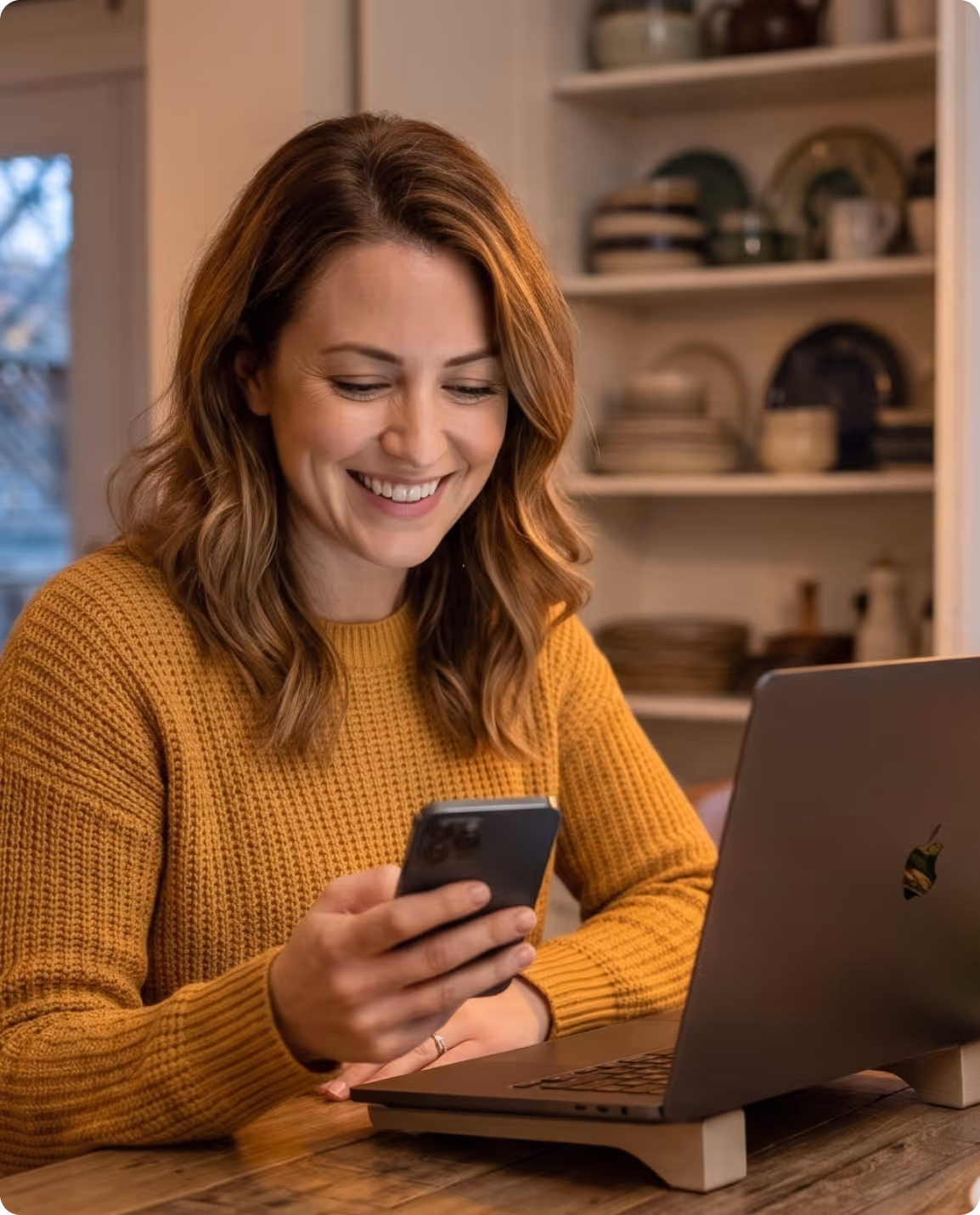 Smiling woman in a mustard sweater looking at her smartphone while sitting at a wooden table with an open laptop.