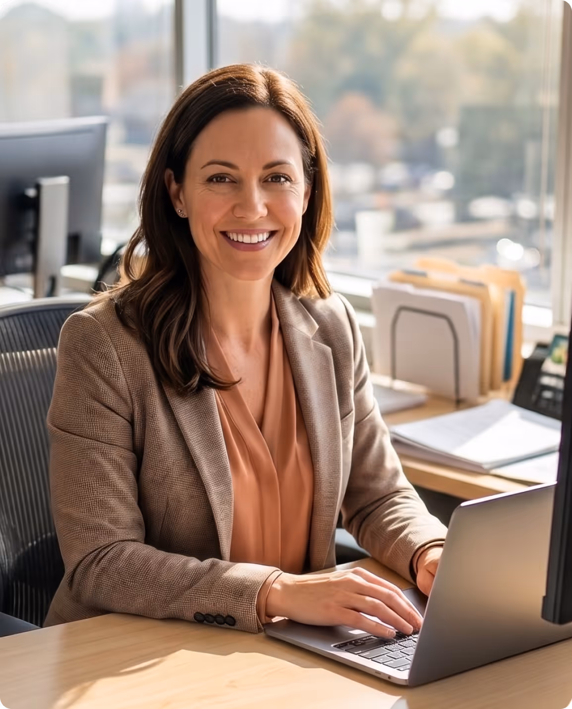 Smiling woman in business attire working on a laptop at an office desk with a window view.