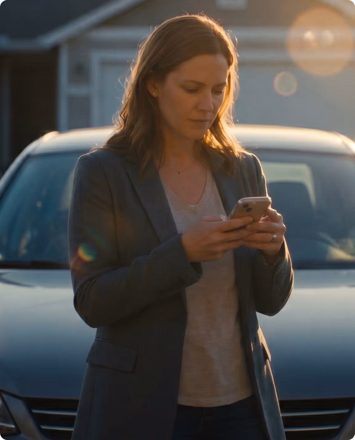Woman in gray blazer standing in front of a car, looking at her phone with sunlight in the background.