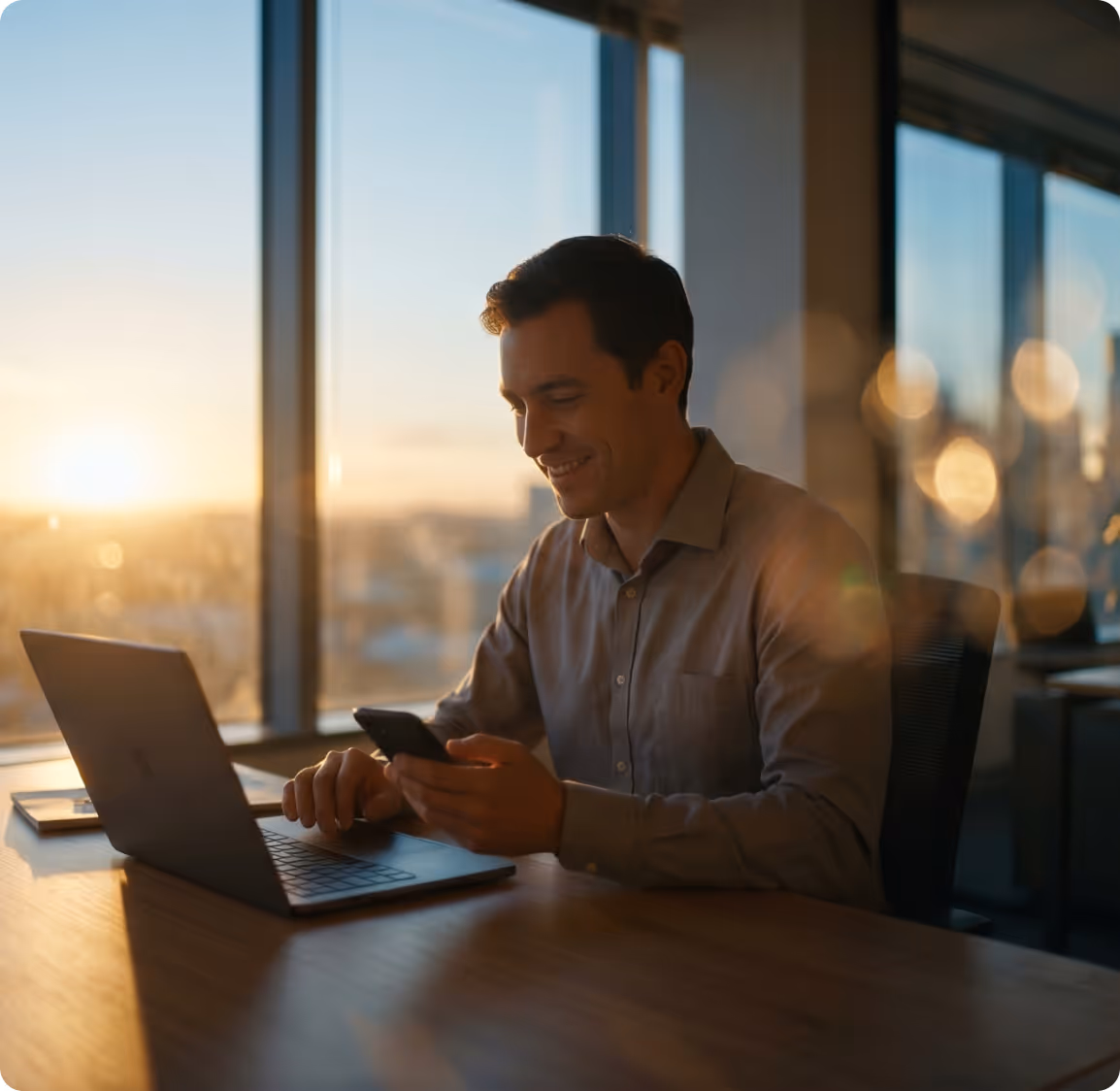 Smiling woman in a light blue shirt using a laptop and holding a smartphone at a wooden desk.