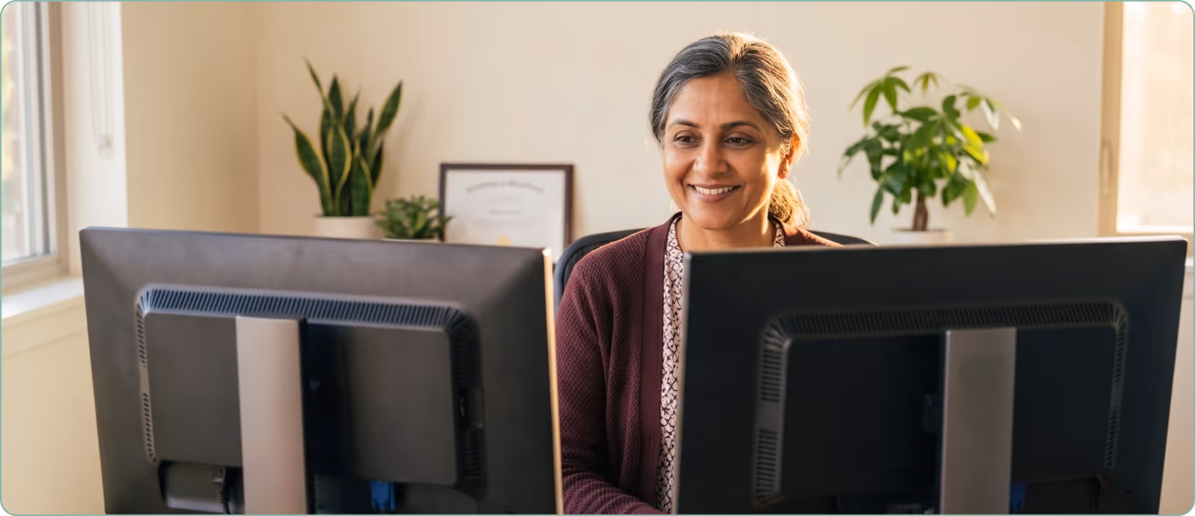 Smiling mature woman with gray hair working on a dual-monitor computer setup in a bright office.