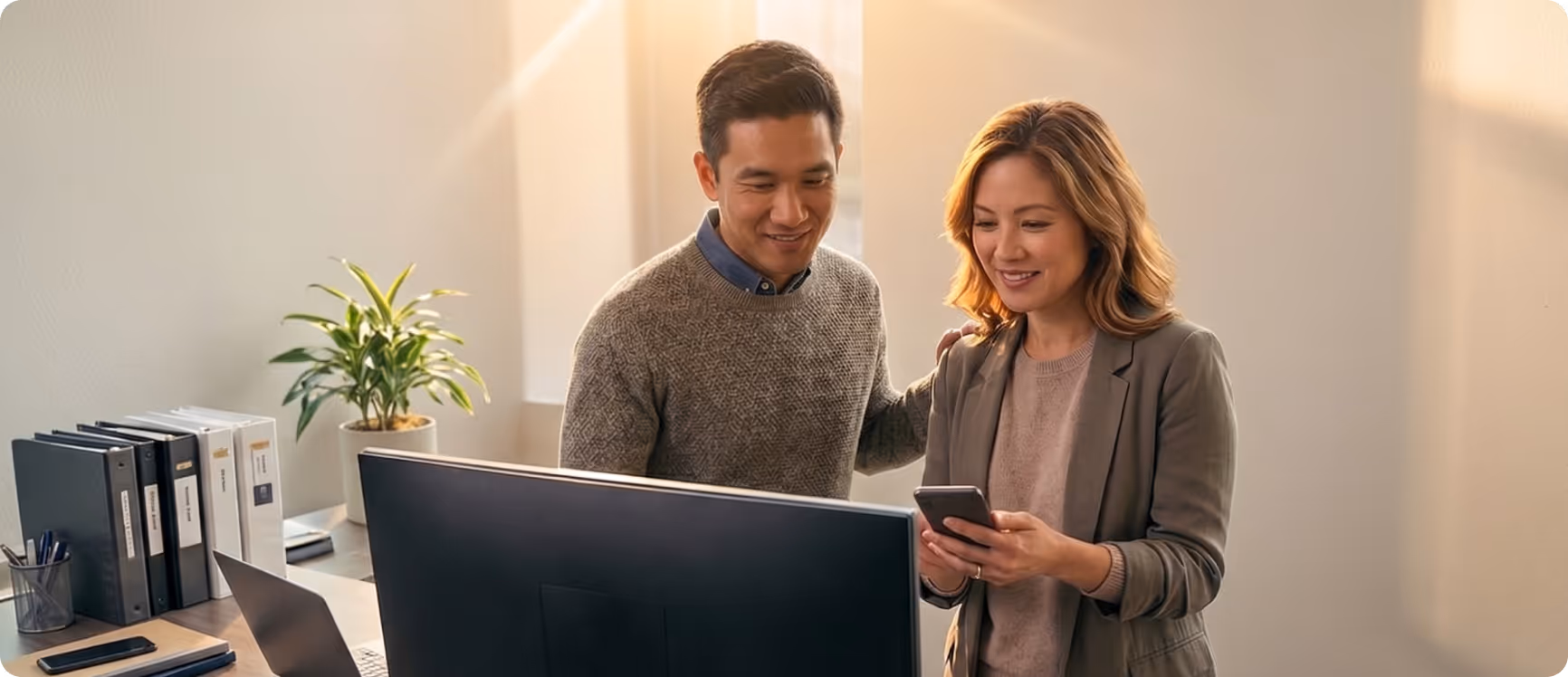 Two colleagues, a man and a woman, smiling and looking at a smartphone together in a bright office.